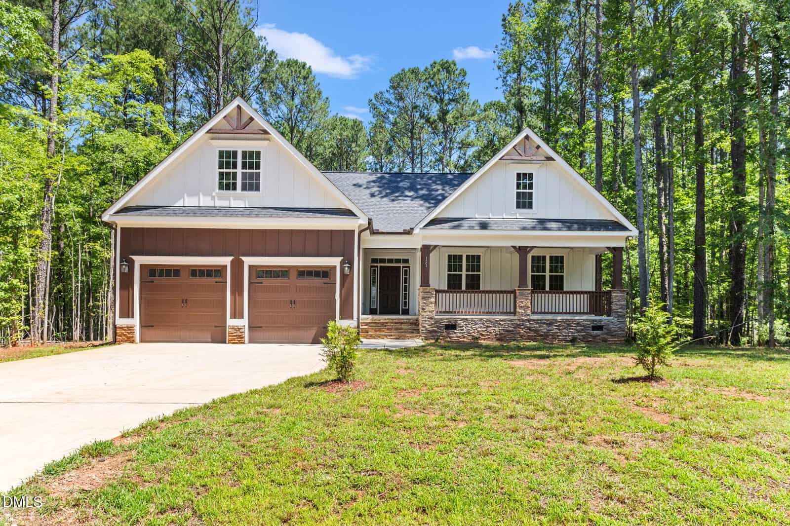 A modern two-story house with a front porch, double garage, and white siding with stone accents, surrounded by tall trees on a bright, sunny day.