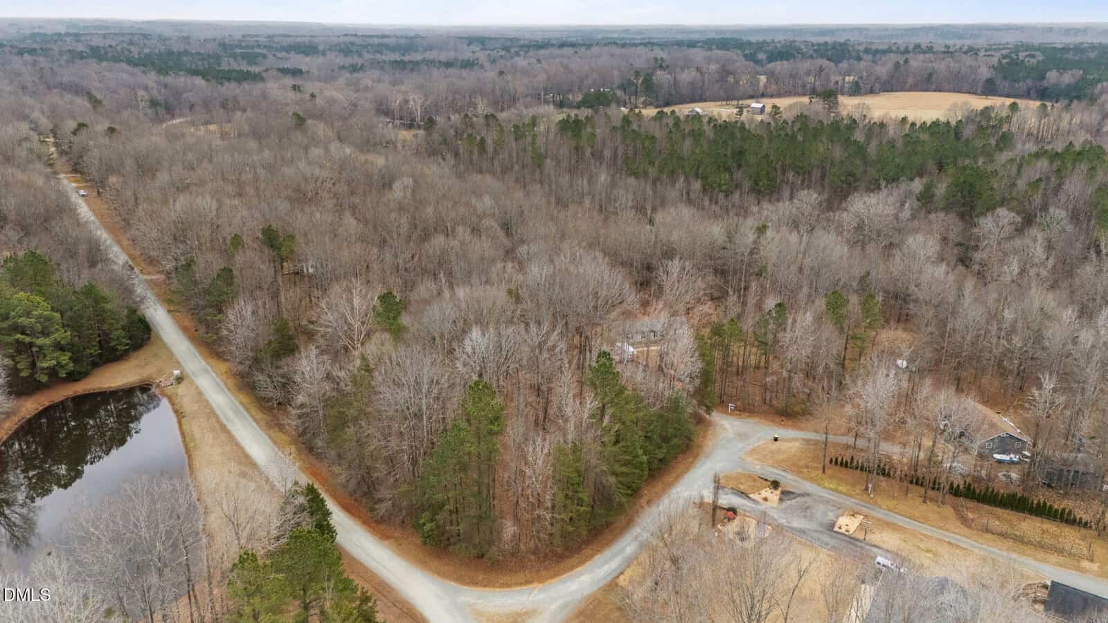 Aerial view of 5590 Shelton Creek Road, Oxford, with intersecting dirt roads, leafless trees, a small pond to the left, scattered houses, and forested land stretching into the distance beneath a cloudy sky.