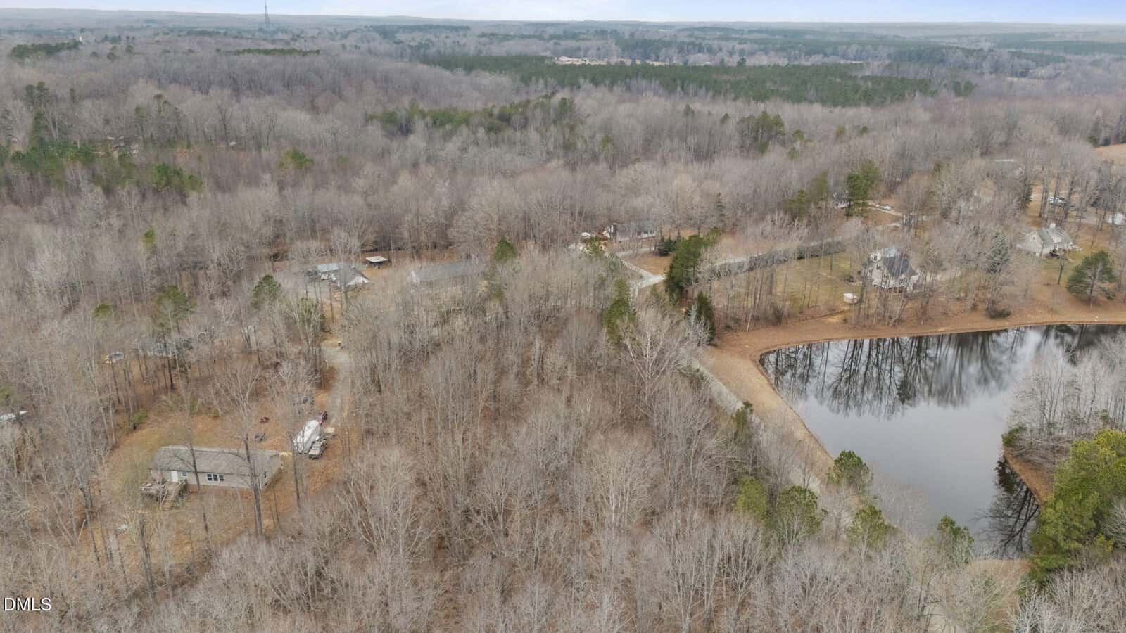 Aerial view of a rural, wooded area with bare trees, scattered houses, a pond with reflections, winding roads, and distant forests under a cloudy sky.