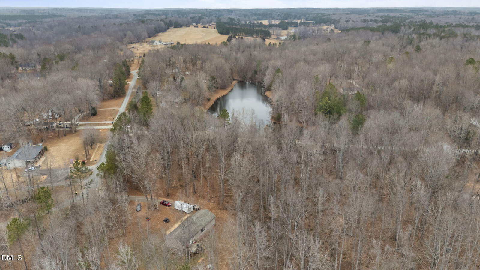 Aerial view of a rural landscape with leafless trees, a small pond in the center, scattered houses and roads, and open fields in the background under a cloudy sky.