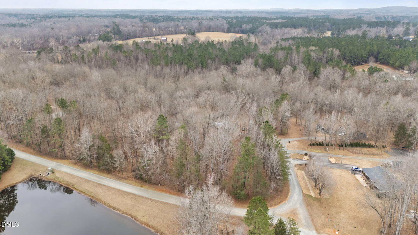 Aerial view of a rural landscape with winding roads, bare trees, a pond, and scattered houses on a cloudy day. The terrain is mostly wooded, with patches of grass and distant rolling hills.