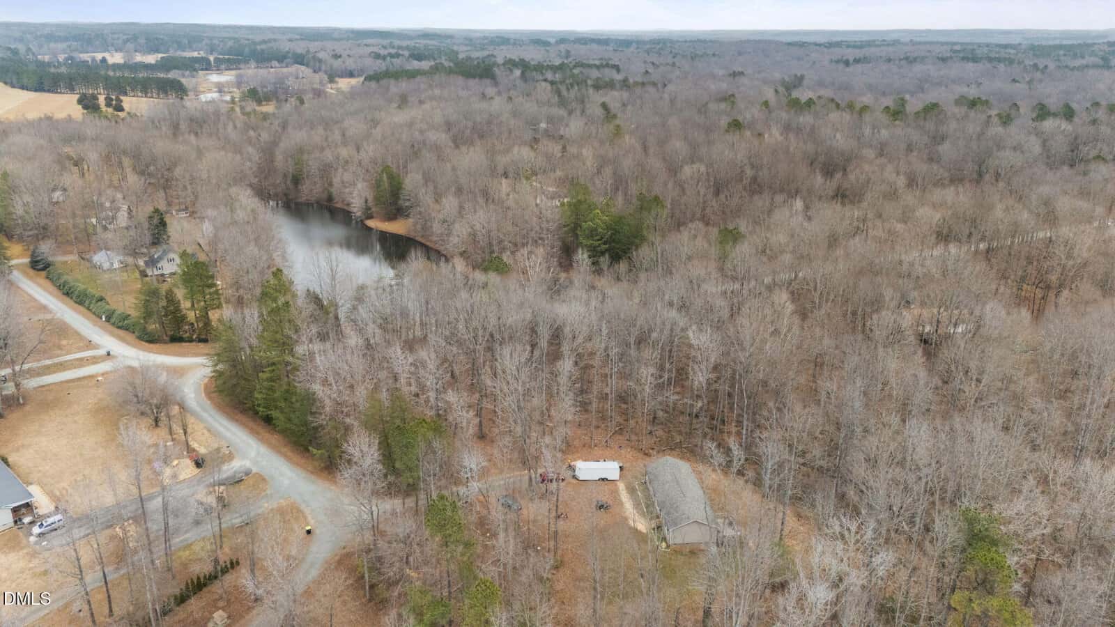 Aerial view of a rural, wooded area with a small pond, scattered houses, a curved road, and leafless trees, likely in late fall or winter. A barn and a white trailer are visible among the trees.