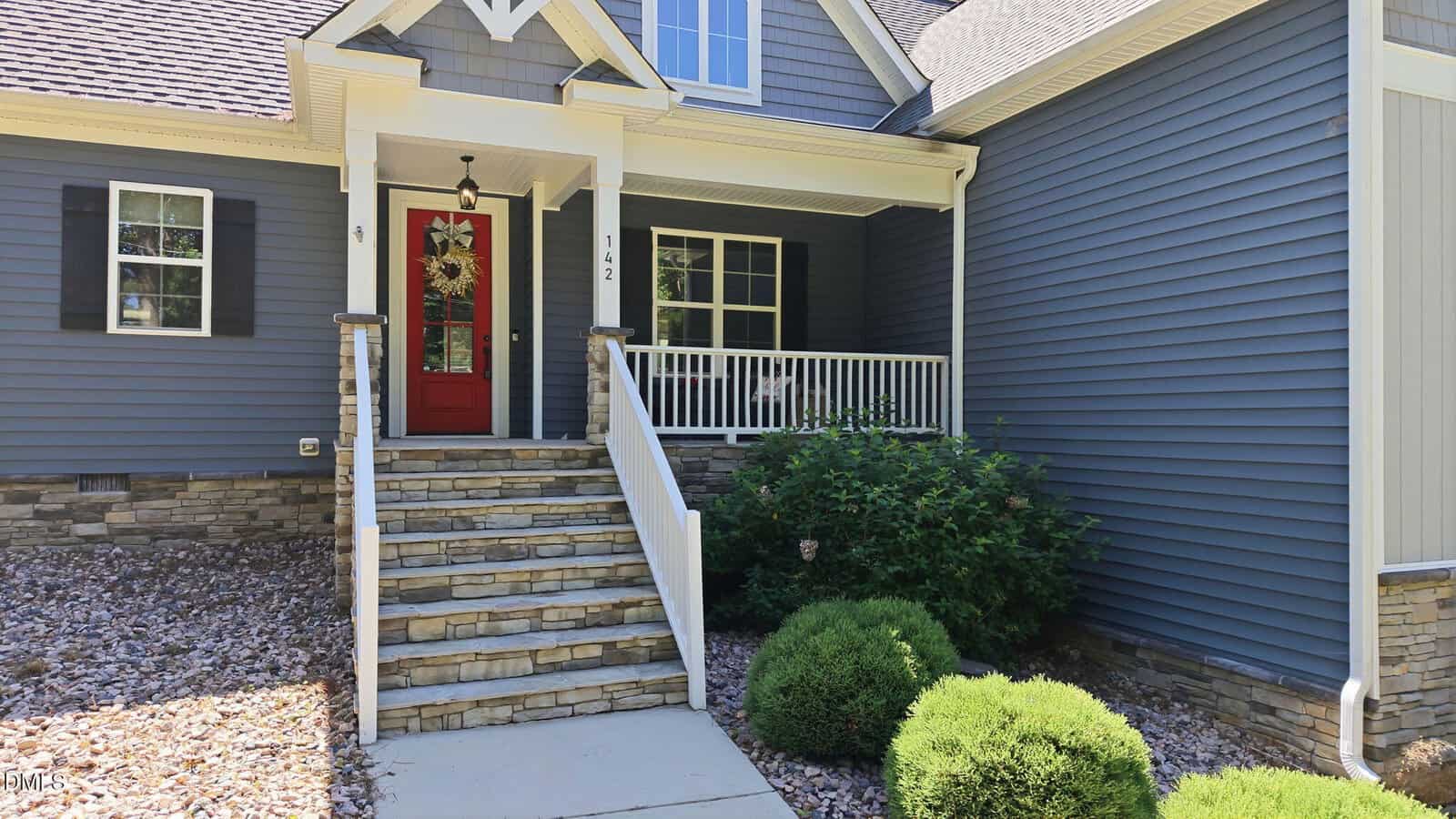 A blue house with stone accents, a covered front porch, white railing, red front door, and a set of stone steps leading to the entry, surrounded by landscaped bushes and rocks.