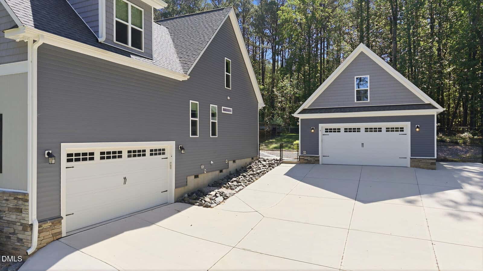 Two gray modern garages with white doors, set on a spacious paved driveway surrounded by trees and stone landscaping, attached to and detached from a house on a sunny day.