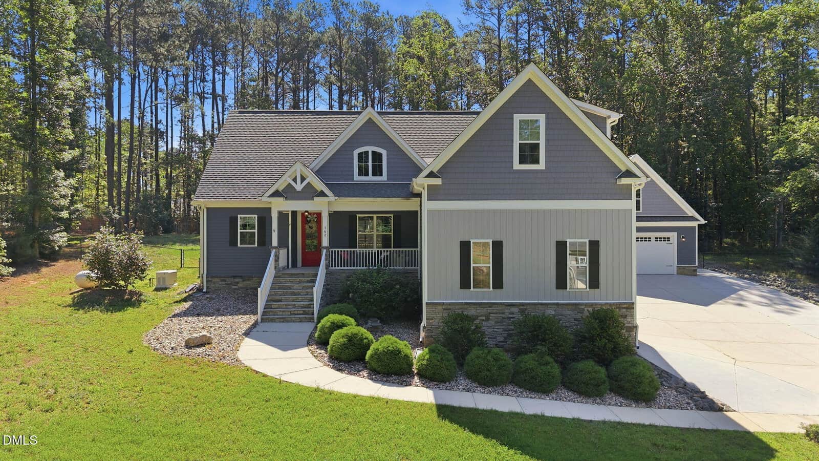Nestled in Louisburg, 142 Mohawk Drive is a gray single-family house with white trim, a front porch, and red door. Set before a wooded area, it features neat landscaping, shrubs along the walkway, and a driveway to a detached garage.