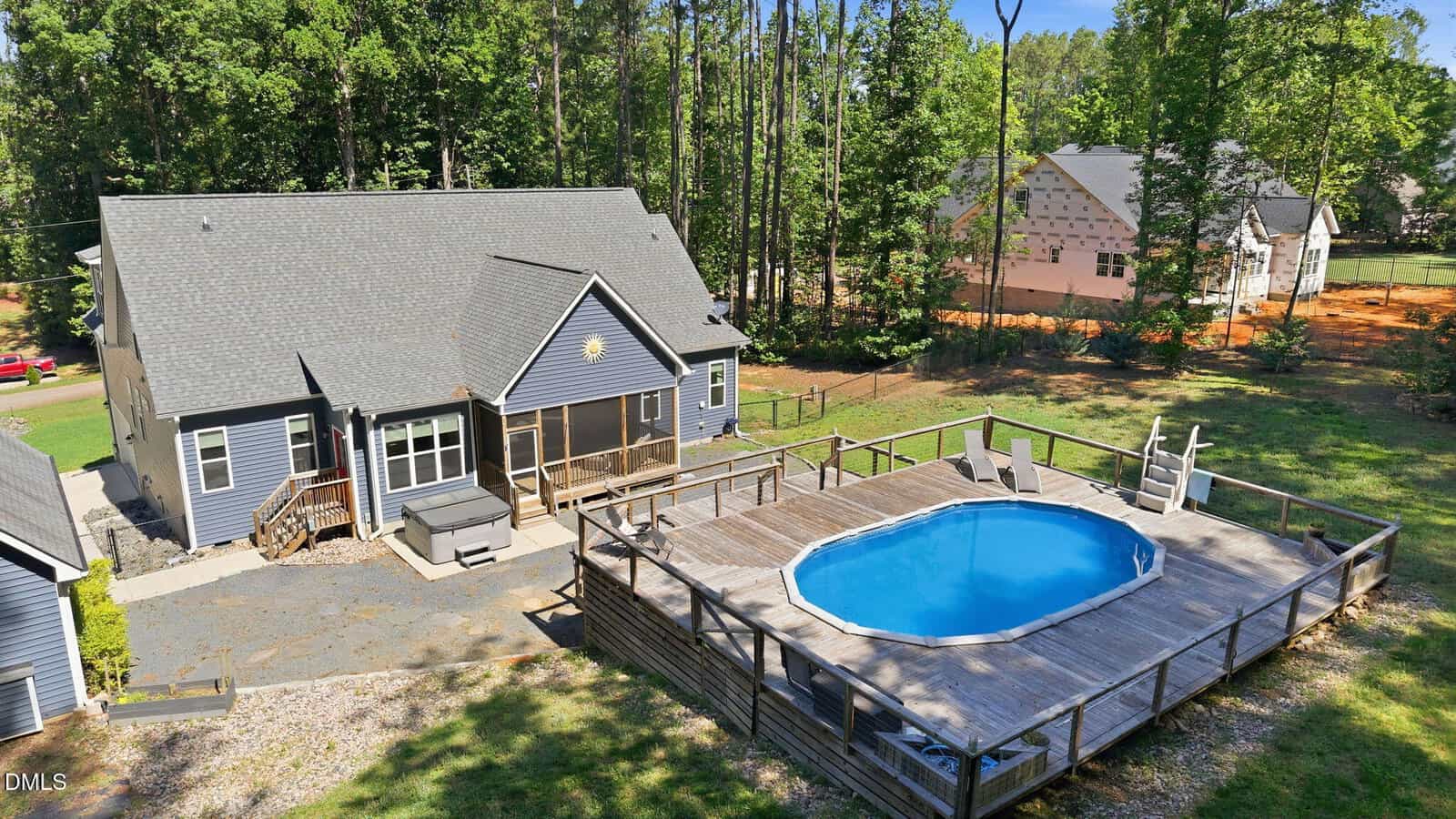 A gray house with a screened porch sits beside a large wooden deck featuring a round above-ground pool, surrounded by trees and neighboring homes in the background.