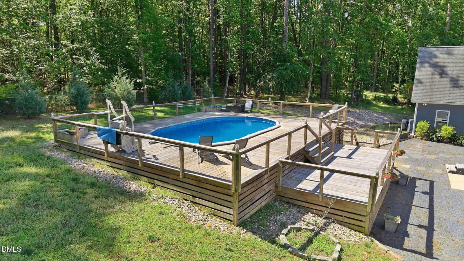 A round above-ground pool with a wooden deck and railing, surrounded by trees. Lounge chairs and small tables are placed on the deck, with steps leading up to the pool area from the ground.