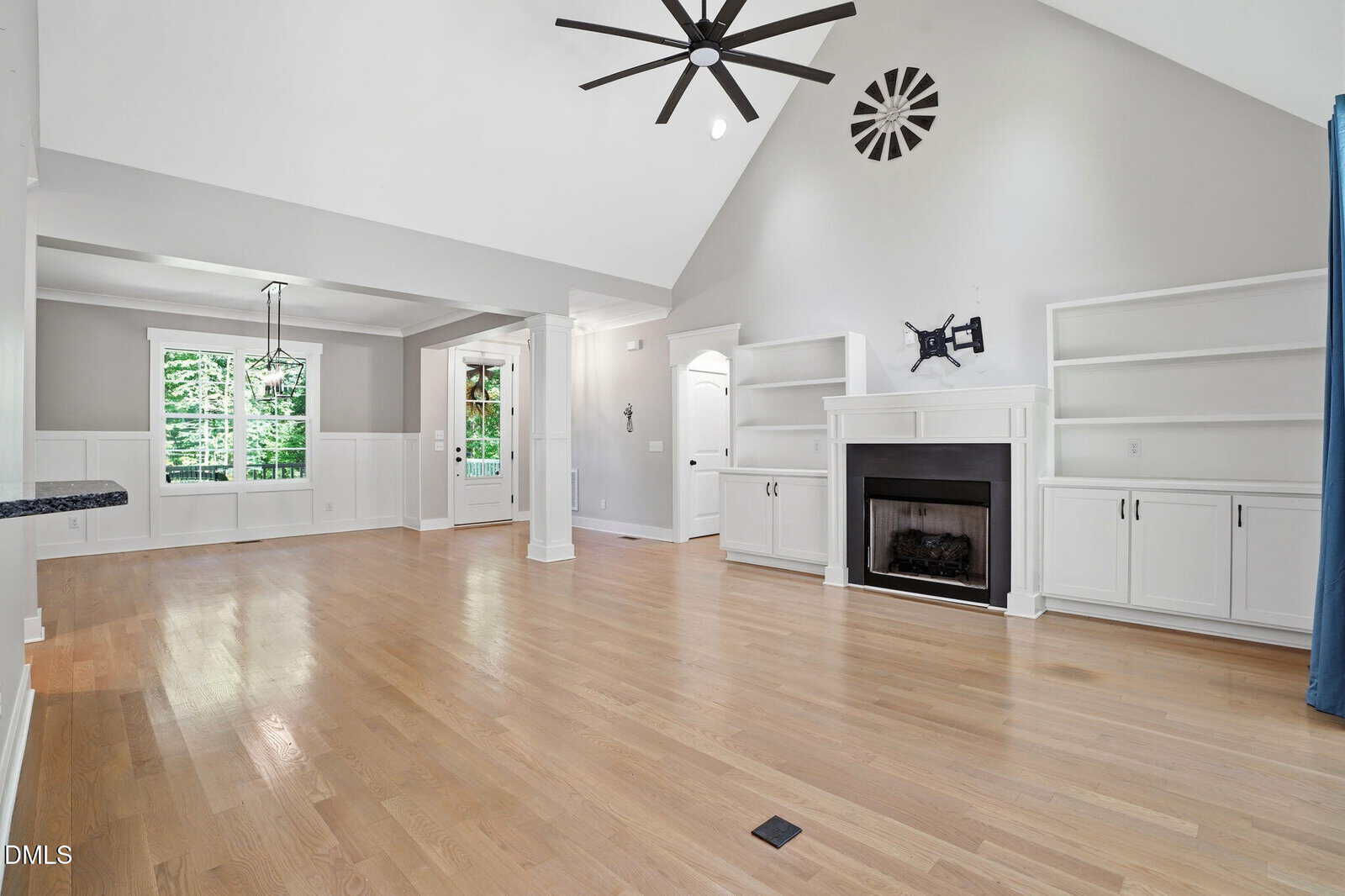 Spacious living room with light wood floors, white built-in shelves, a fireplace, vaulted ceiling, modern fan, and large windows letting in natural light. Dining nook is also visible.