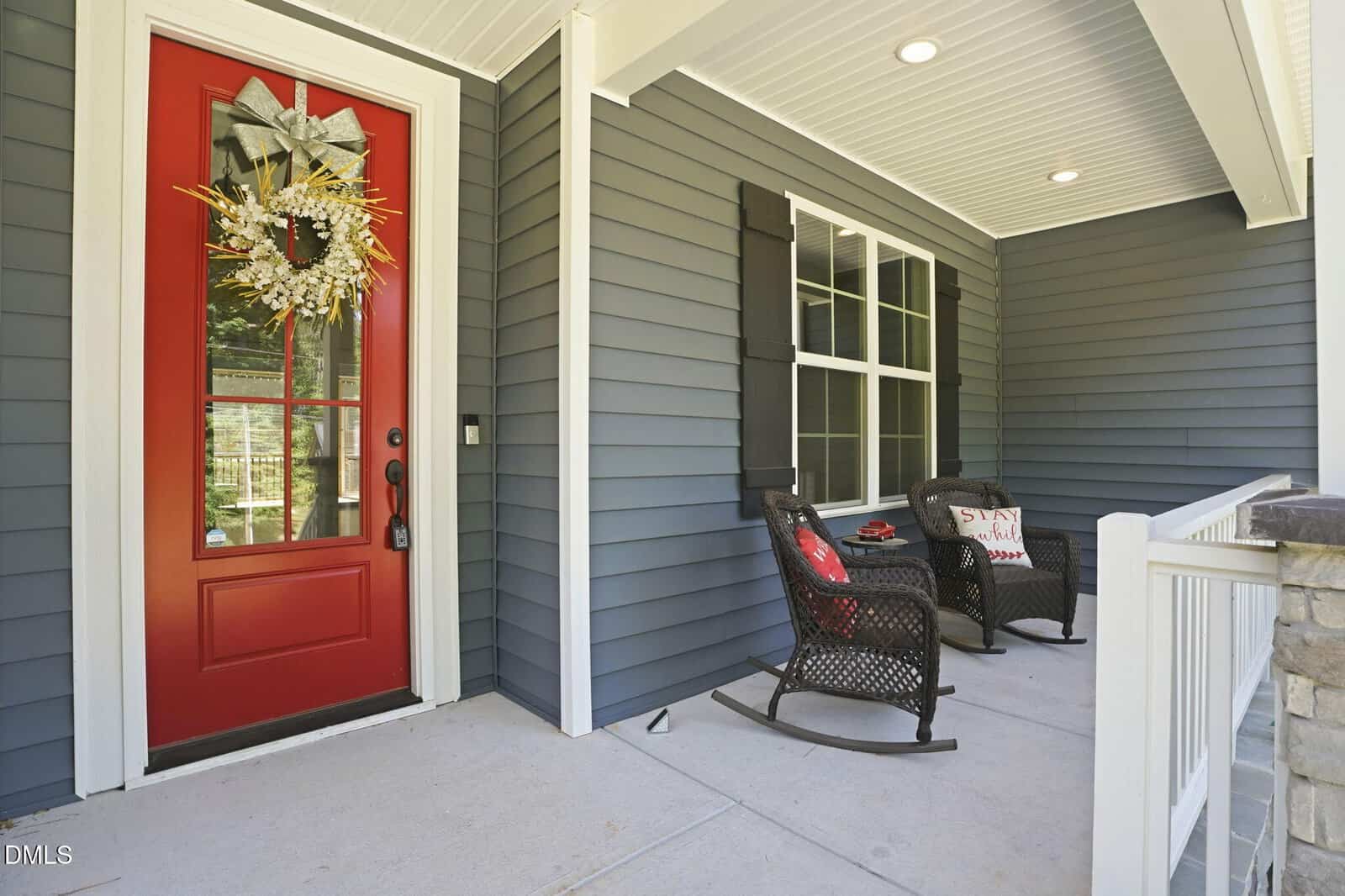 A front porch with gray siding, a red door decorated with a wreath, two black rocking chairs with cushions, and a small table. The porch has white trim and a covered ceiling with recessed lights.