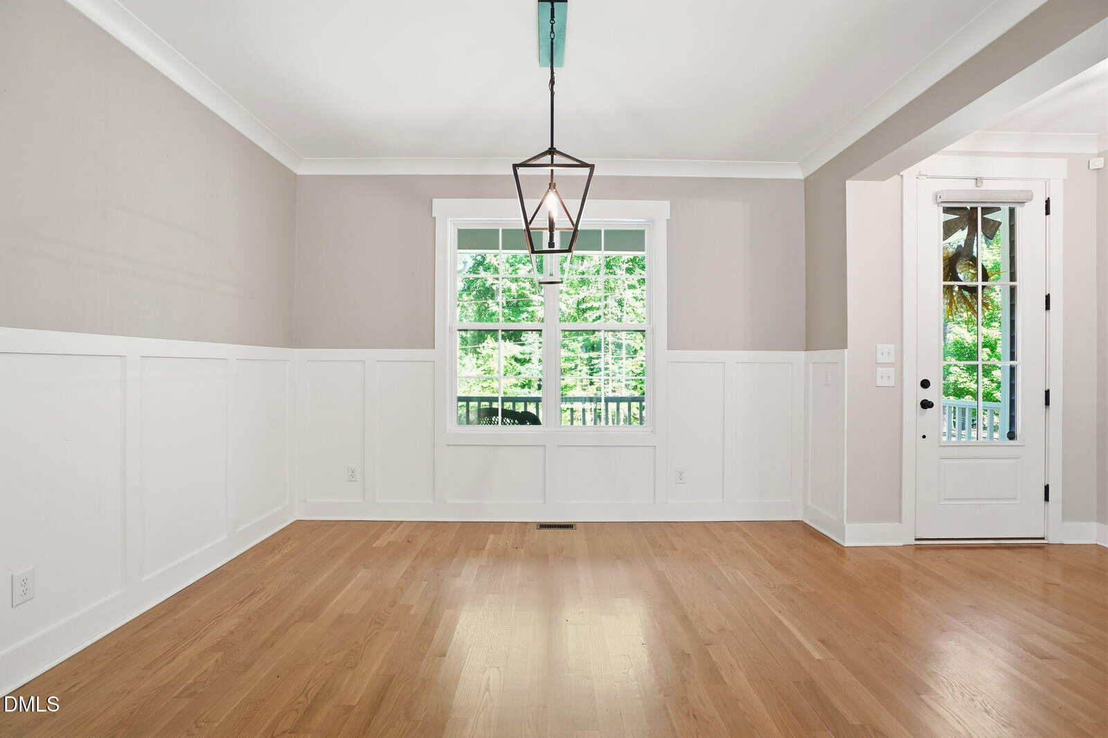 Bright, empty dining room with light wood floors, white wainscoting, light gray upper walls, large window, geometric pendant light, and a door to the outside.