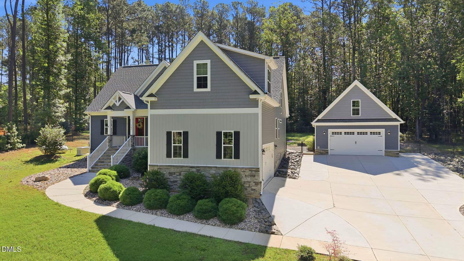 Gray two-story house with stone accents, front porch, and red door, situated on a landscaped yard with shrubs. Separate two-car garage in the background, surrounded by trees and a large concrete driveway.