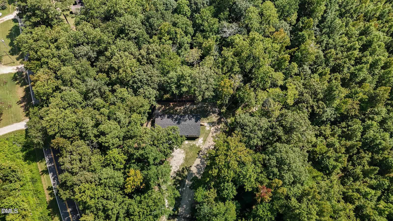 Aerial view of a small house surrounded by dense green trees and forest, with a driveway leading to the house from a nearby road on the left side of the image.