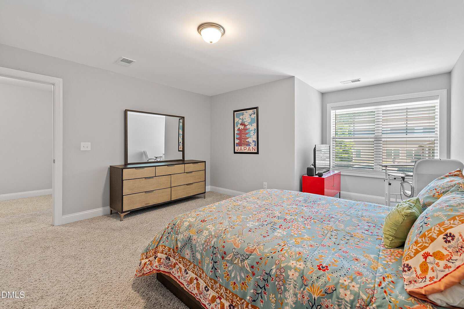 A bright bedroom with a bed featuring a colorful floral comforter, a wooden dresser with a mirror, a framed Japan travel poster, and a desk by a window with blinds allowing natural light in.
