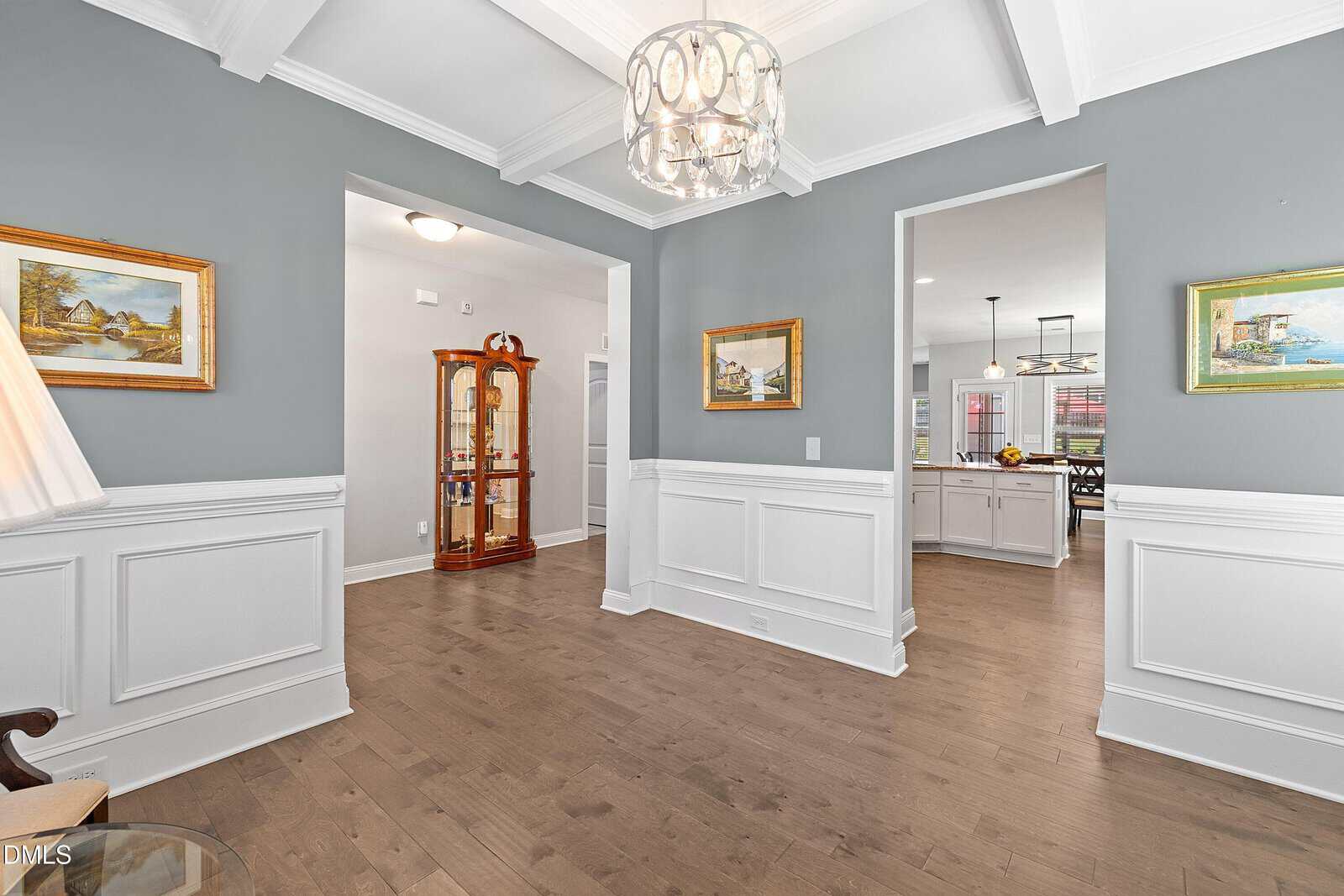 A bright, elegant room with gray walls, white trim, wood floors, and crown molding. Framed art hangs on the walls, and a chandelier hangs from the coffered ceiling. A kitchen is visible through an open doorway.