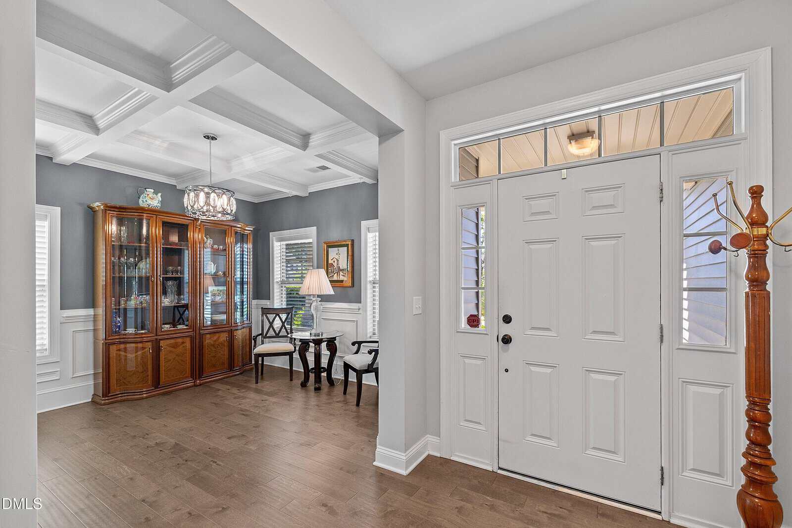 Bright entryway with double doors, sidelights, and a coat rack. To the left is a dining area with a wooden china cabinet, two chairs, a table, and a floor lamp, featuring gray walls and white trim.