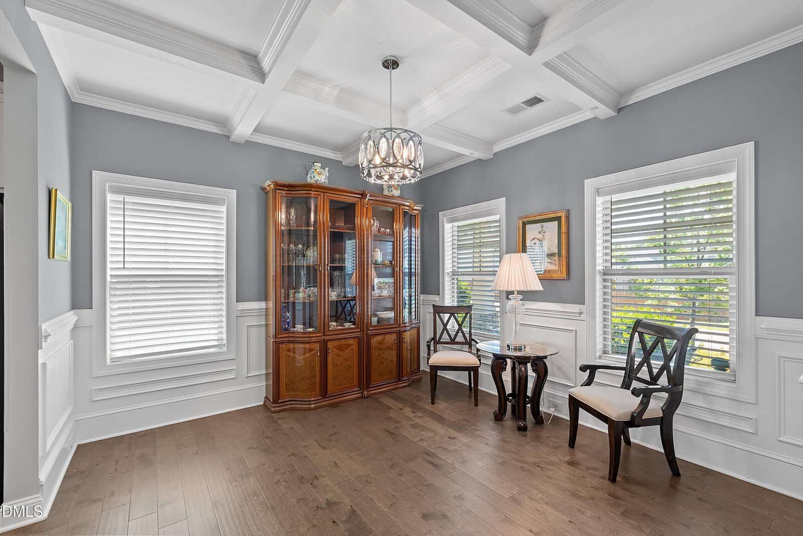 A bright sitting room with gray walls, wood flooring, a glass-front china cabinet, two chairs, a small table with a lamp, and a modern chandelier. Sunlight streams through white-shuttered windows.