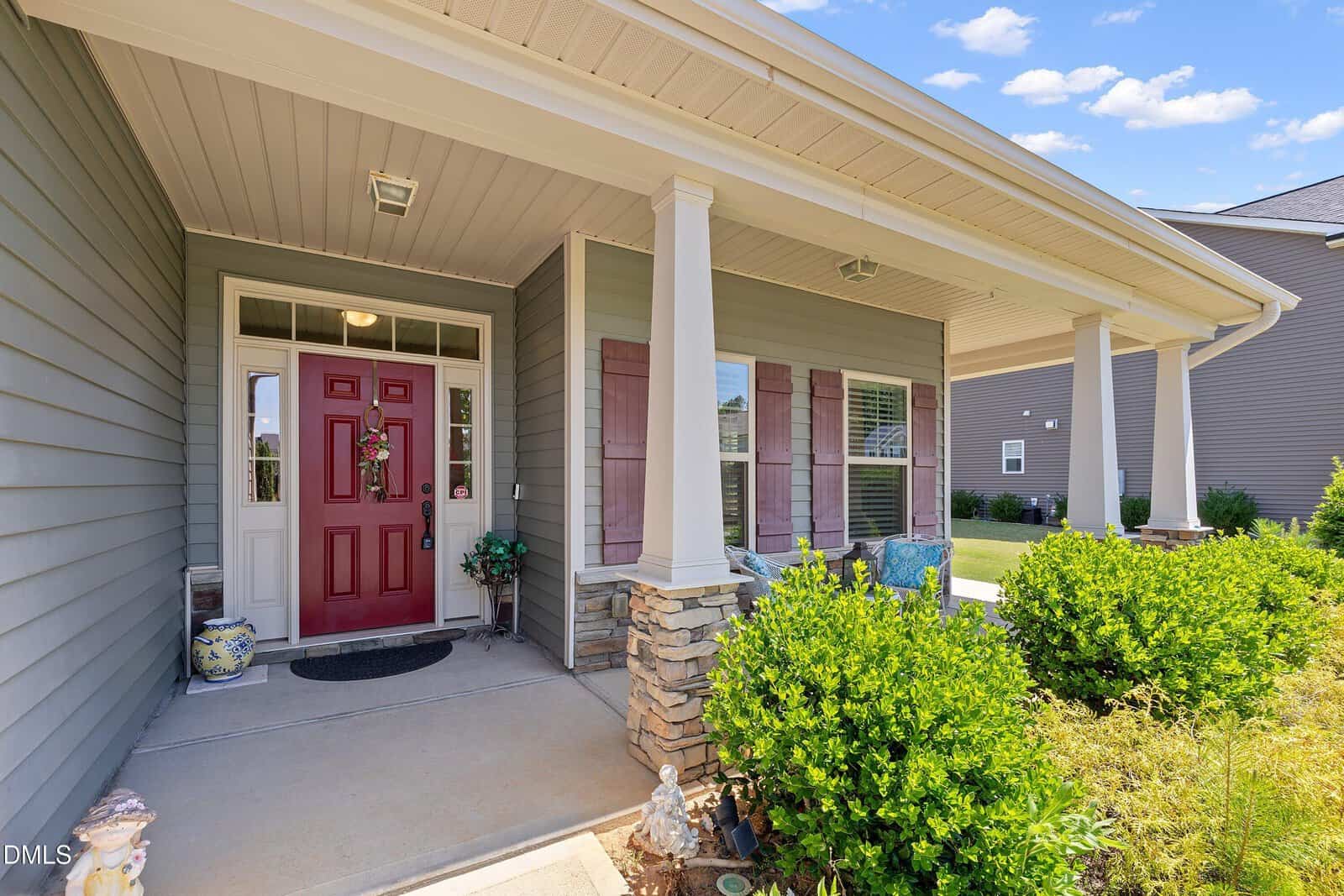 A front porch with white columns, stone accents, and a red door. Shrubs and greenery line the walkway, and there are decorative items near the entrance. The exterior walls are gray with maroon shutters.
