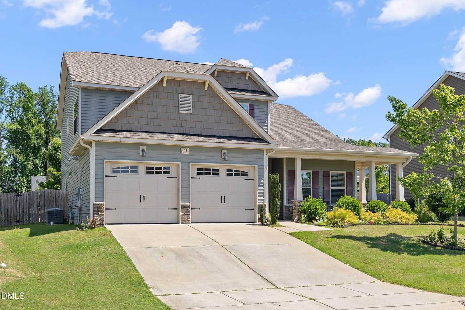 Two-story suburban house with a double garage, gray siding, and a covered front porch. Green lawn and bushes surround the entrance, with a blue sky and scattered clouds overhead.