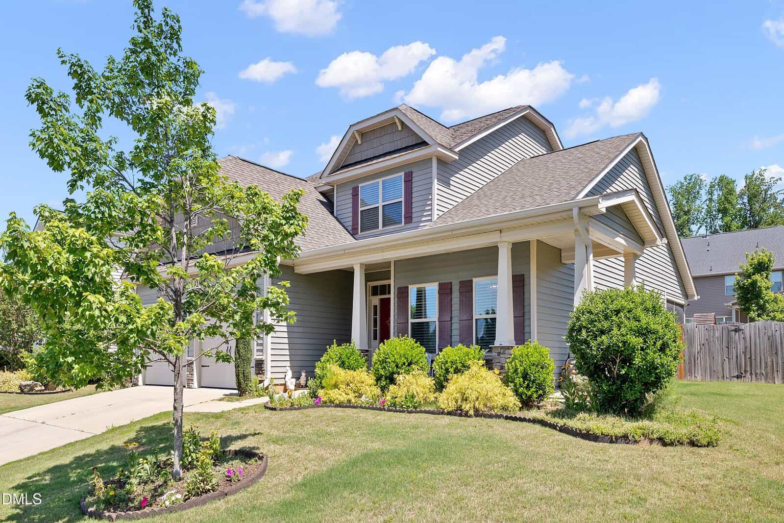 A modern two-story house with gray siding, a front porch, manicured lawn, shrubs, and a young tree, on a sunny day with blue sky and scattered clouds.
