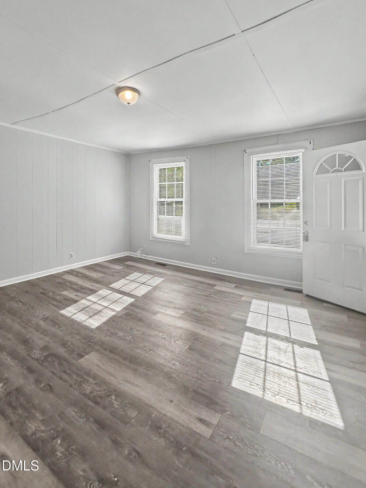 A bright, empty room with gray wood flooring, light gray walls, two windows, and an open white door letting in sunlight that casts window-shaped shadows on the floor.