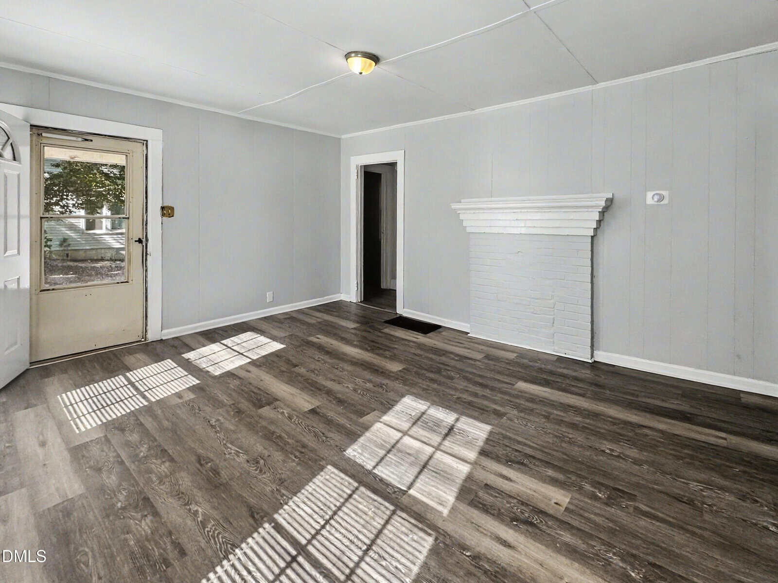 Empty living room with gray walls, white trim, vinyl wood flooring, a white brick fireplace, and a window casting shadows on the floor. A door to the outside and two interior doorways are visible.
