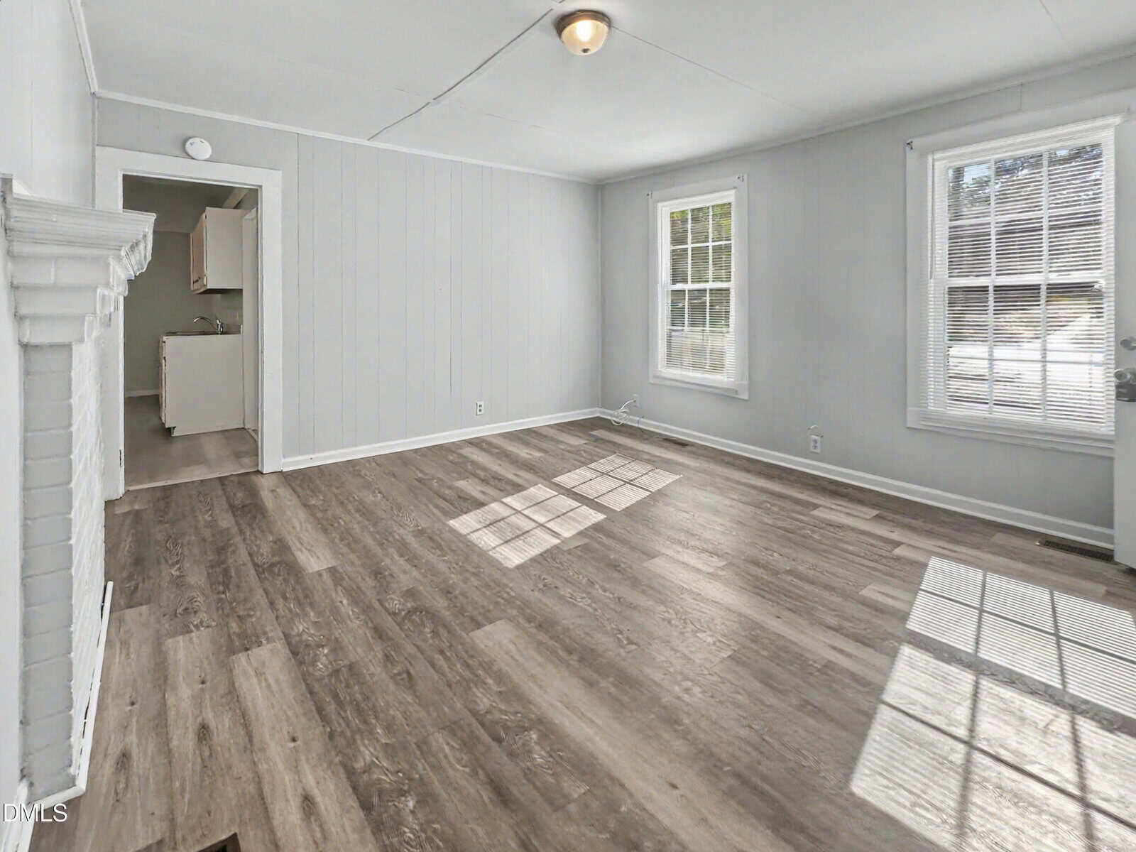 A bright, empty living room with light gray walls, two large windows, wood-style flooring, and a view into a small adjoining kitchen. Sunlight casts shadows across the floor.