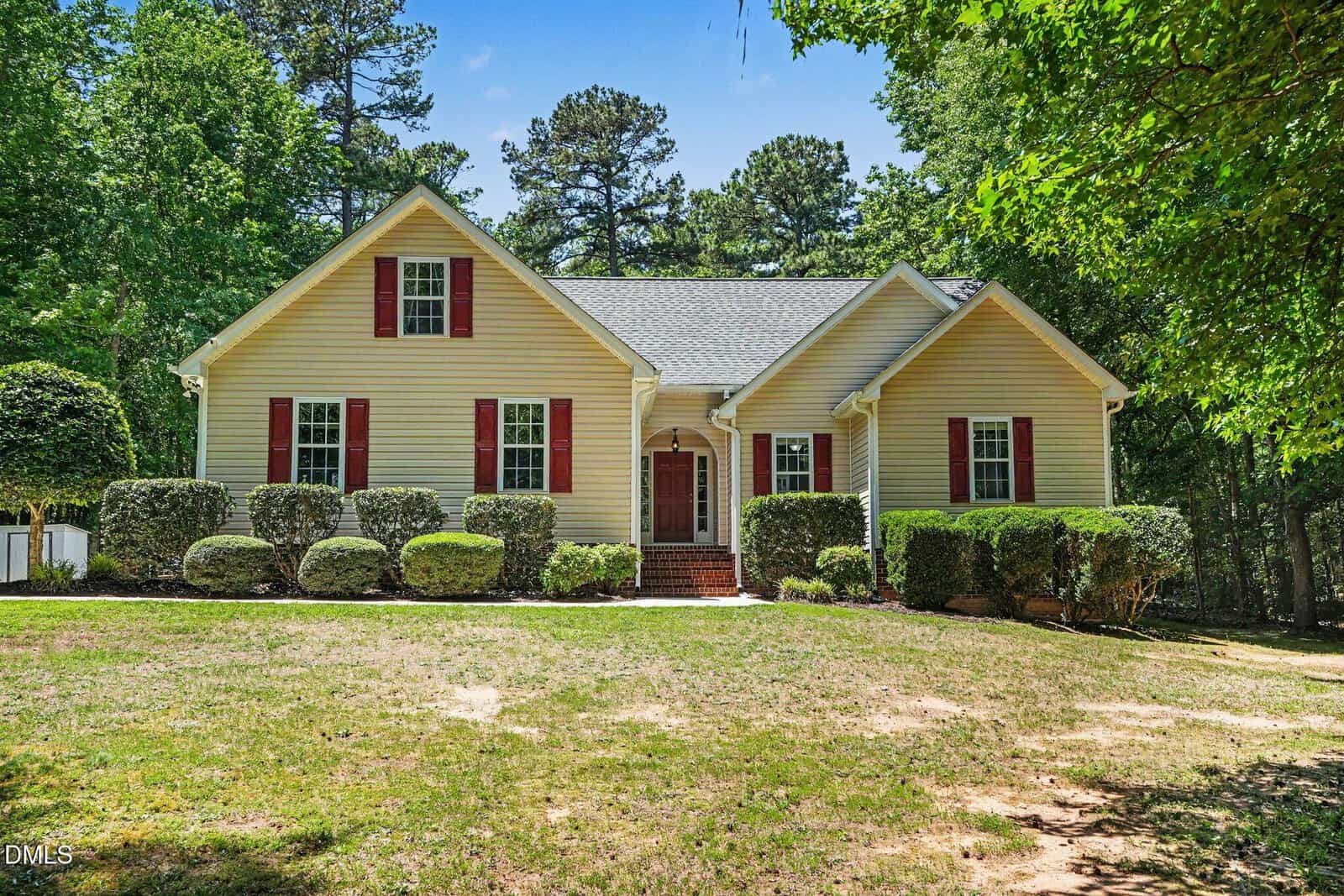 A beige, single-story house at 195 Lumberjack Lane in Louisburg with red shutters and a central front door, surrounded by neatly trimmed bushes and trees. The home sits on a grassy lawn with a wooded backdrop under a blue sky.
