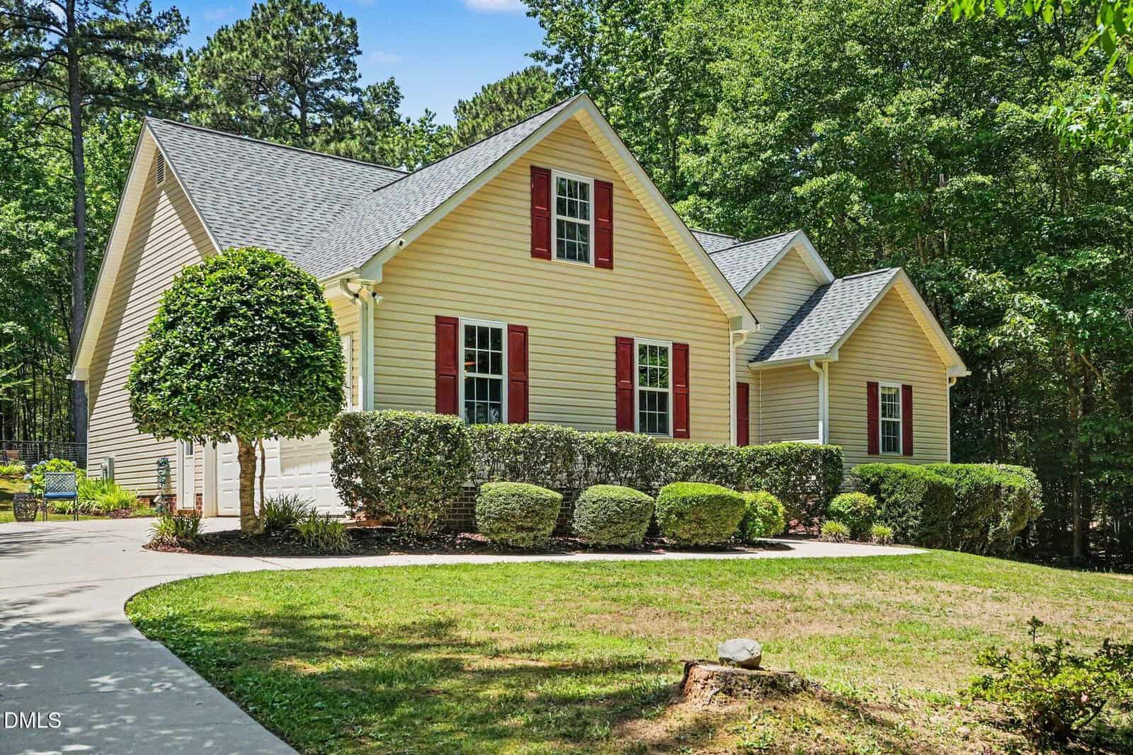 A yellow house with red shutters and white trim, surrounded by neatly trimmed shrubs and trees, sits next to a curved driveway and a wooded area in the background.