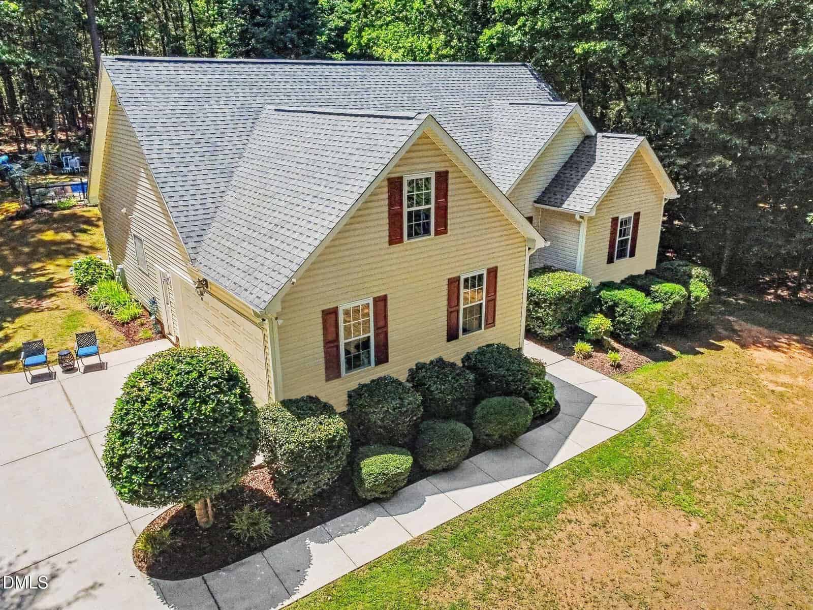 A two-story beige house with a gray shingle roof, red window shutters, landscaped bushes, and a concrete driveway, surrounded by trees and a grassy lawn.