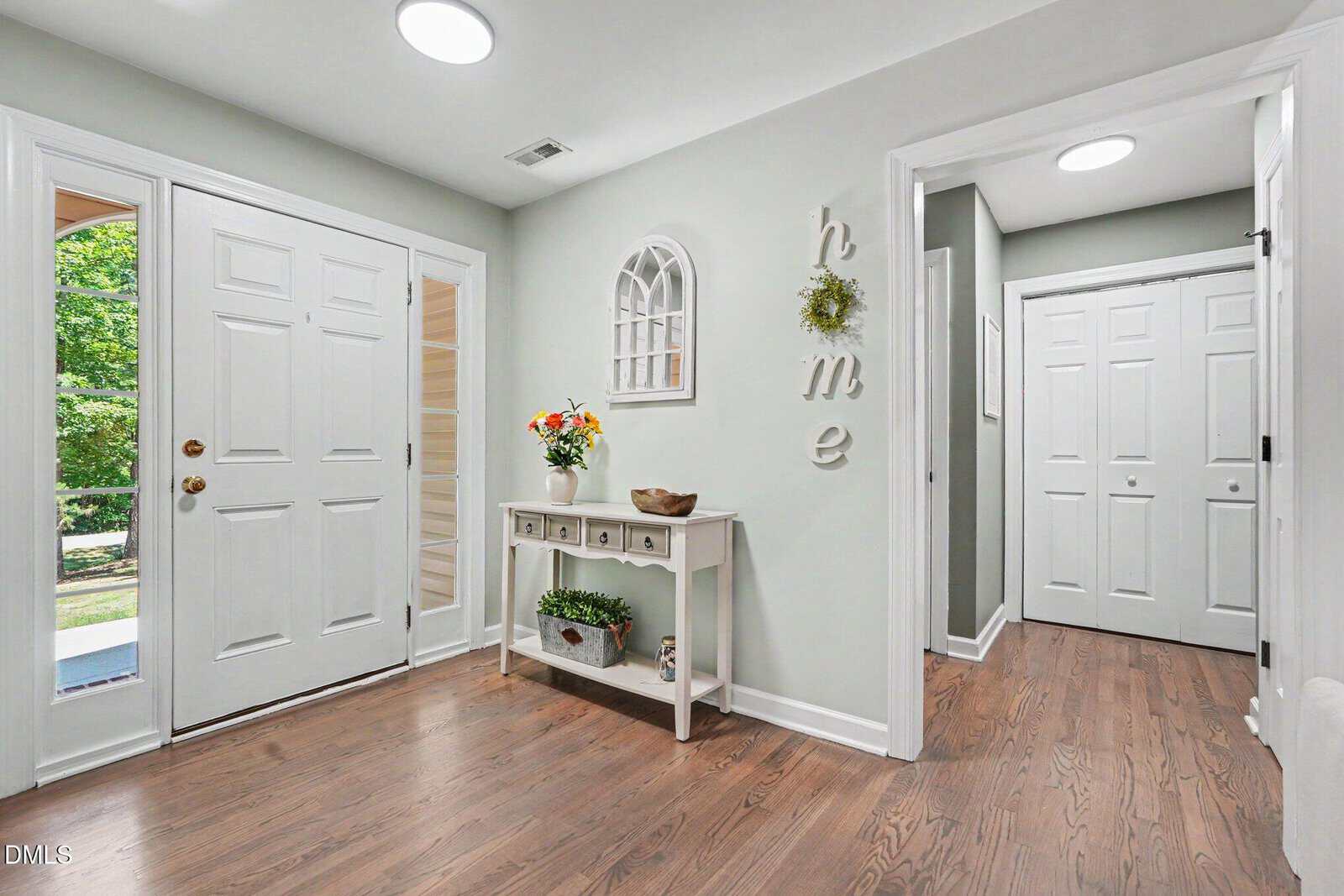 Bright entryway with wood floors, light gray walls, and white trim. A white console table with drawers holds flowers and decor. "Home" decor letters hang on the wall. Doors and hallways lead to other rooms.