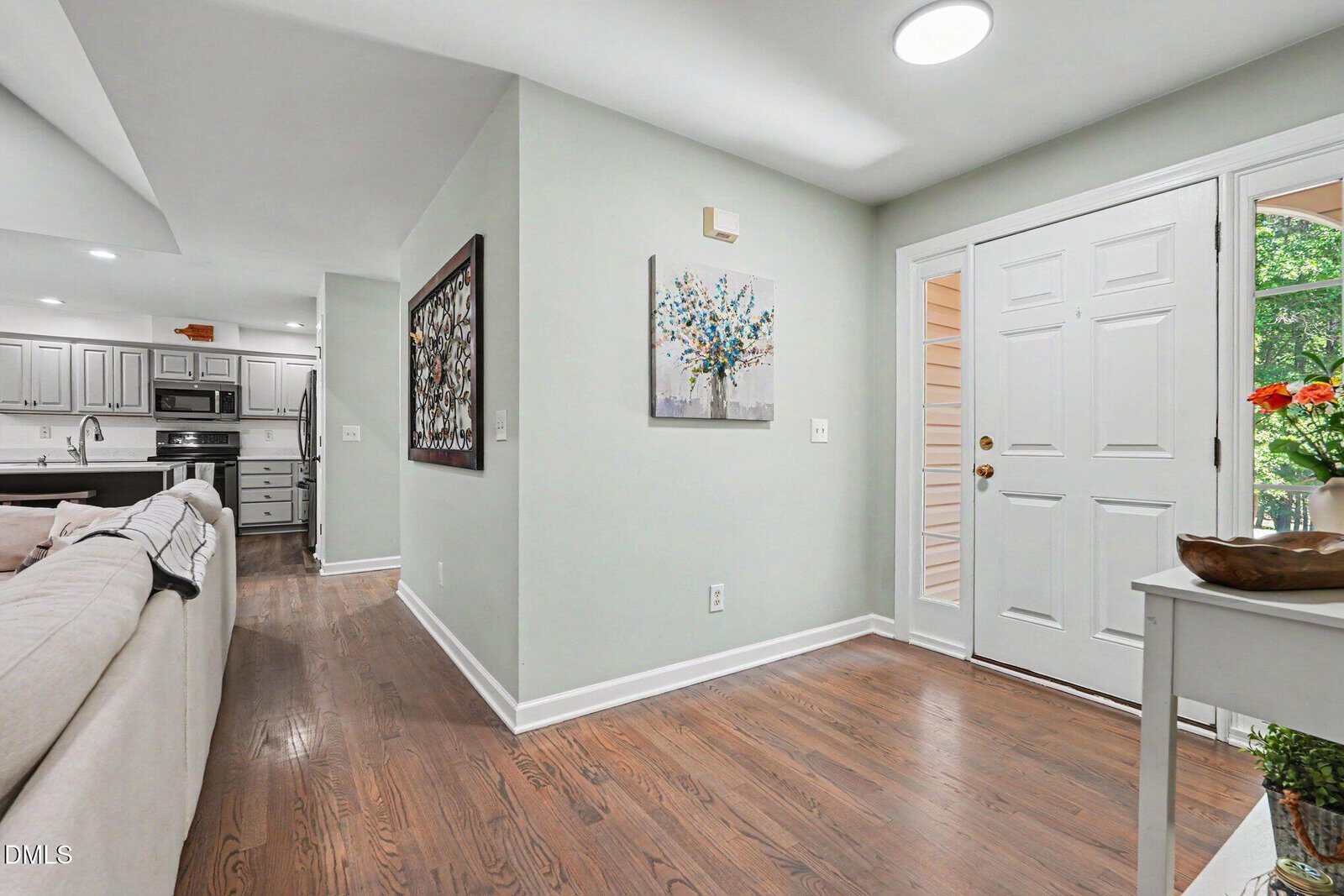 A bright entryway with light green walls, a white front door, hardwood floors, and a small white table with flowers. The entry opens to a living area and kitchen with white cabinetry and stainless steel appliances.