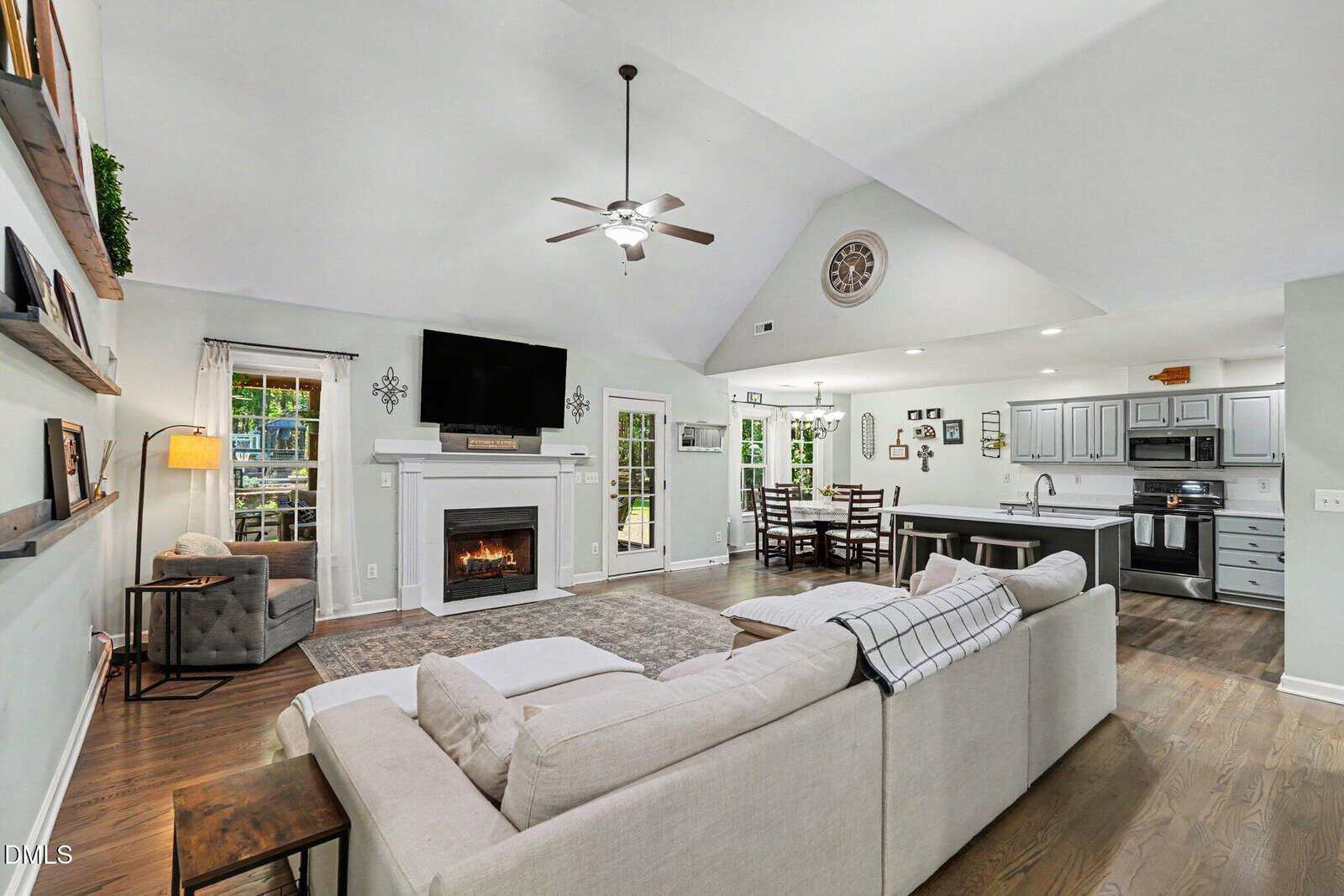 Spacious open-concept living room and kitchen with vaulted ceiling, light gray sofa, wall-mounted TV above fireplace, dining area, and modern gray cabinetry. Large windows let in natural light.