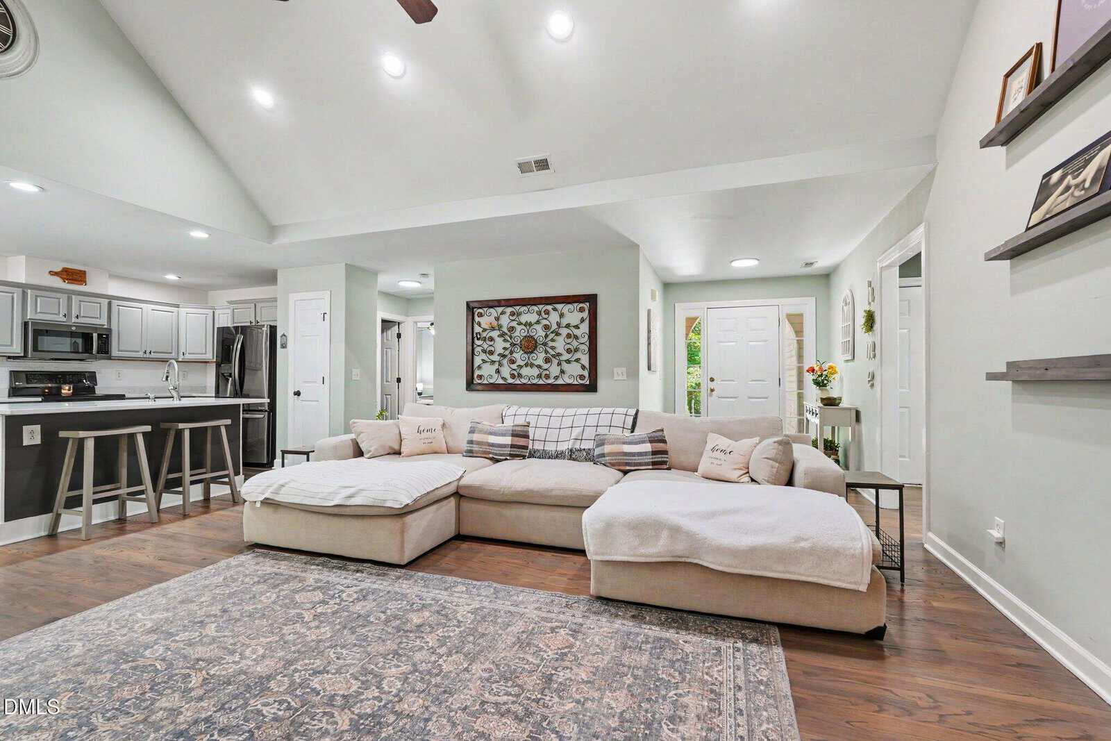 Modern living room with a large beige sectional sofa, decorative pillows, a patterned rug, and an open kitchen in the background. Walls are light-colored, and recessed lighting brightens the space.