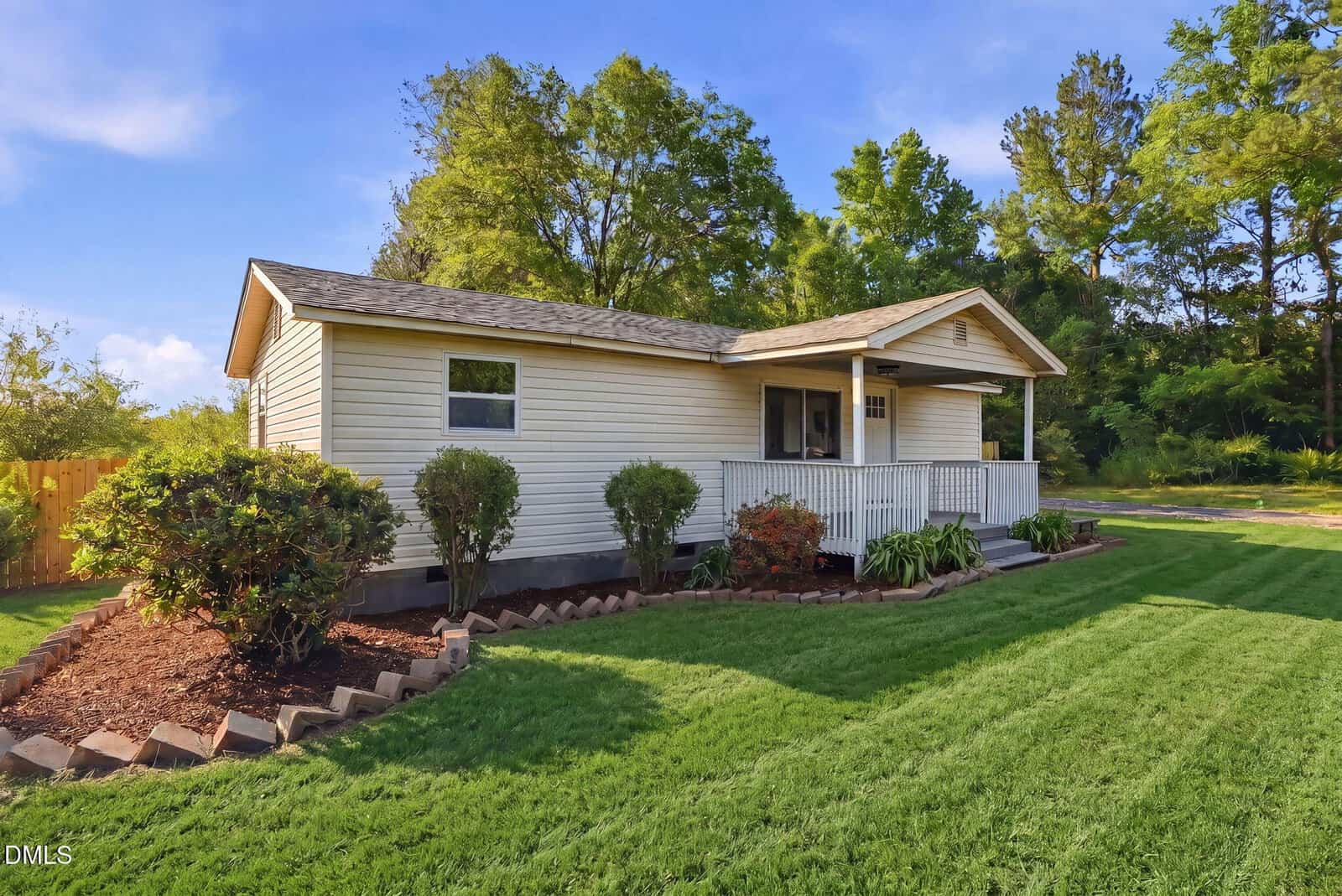 A small, single-story beige house at 1795 Hicks Road, Youngsville, features a front porch with white railing, neatly trimmed shrubs, a well-maintained lawn, and is surrounded by trees under a blue sky.