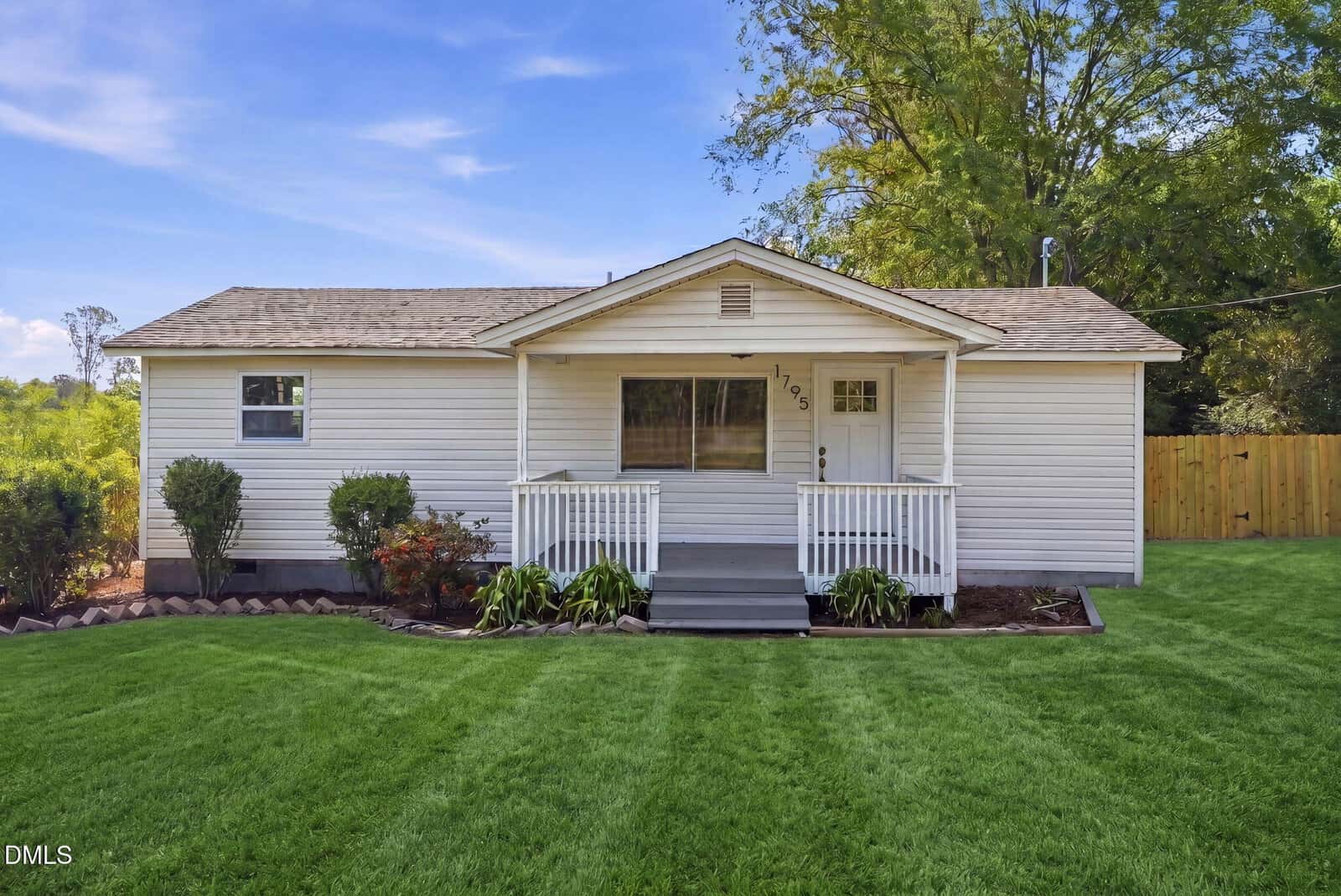 A small, single-story white house with a covered front porch, steps, and railing, surrounded by a well-manicured green lawn and some shrubs, with trees and a wooden fence in the background.