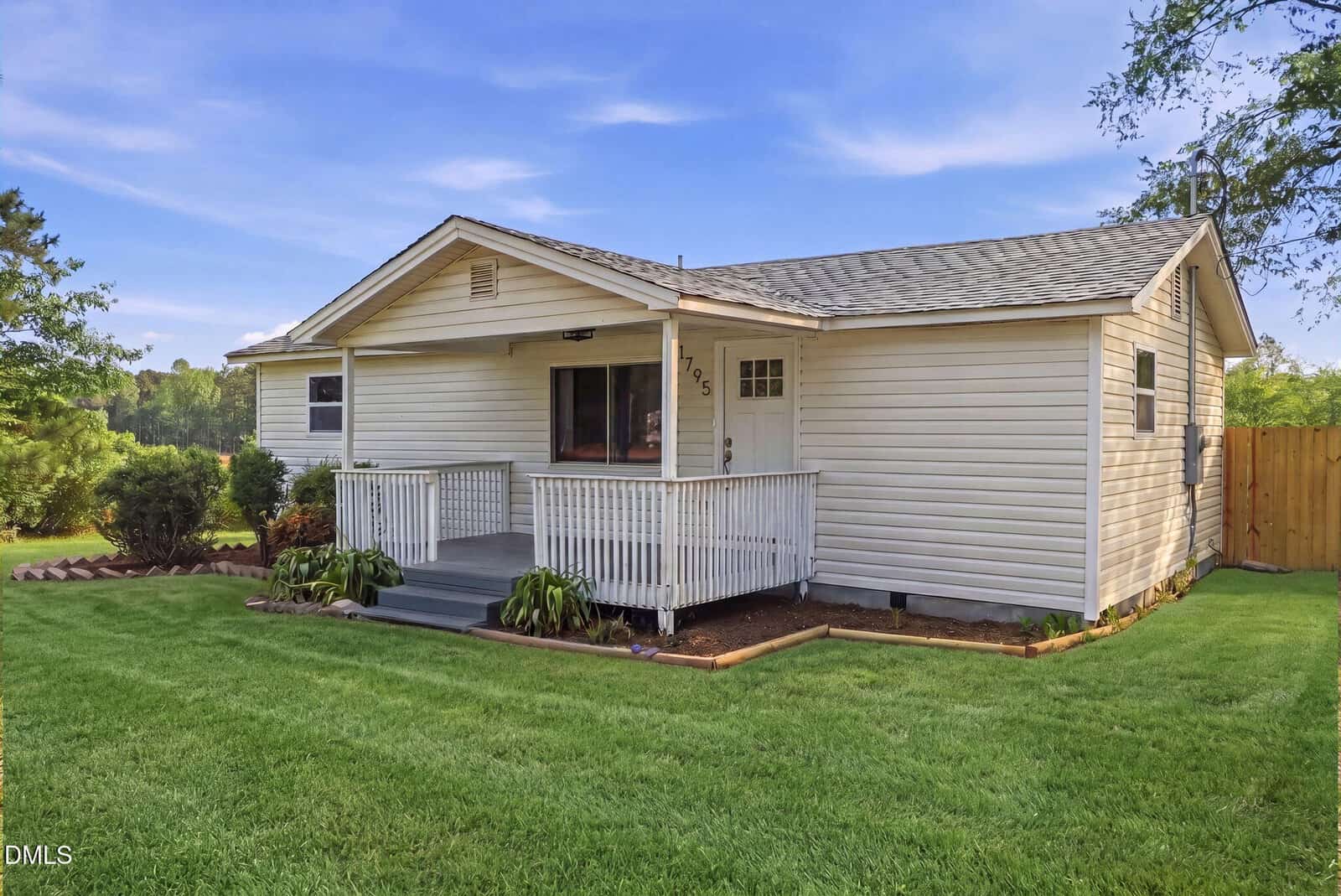 A small, single-story house with beige siding and a covered front porch. The porch has white railings and steps leading to a well-kept green lawn. Trees and a wooden fence are visible in the background.