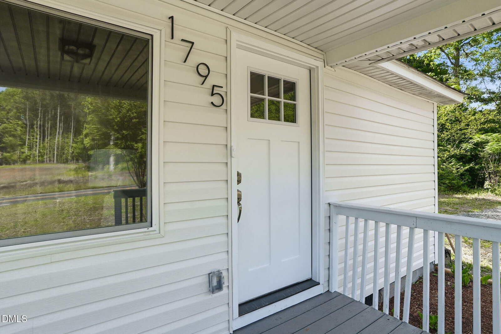 White front door with four small glass panes, black house number 1795 next to it, light gray siding, white railing, and a green wooded area reflected in the window and visible in the background.