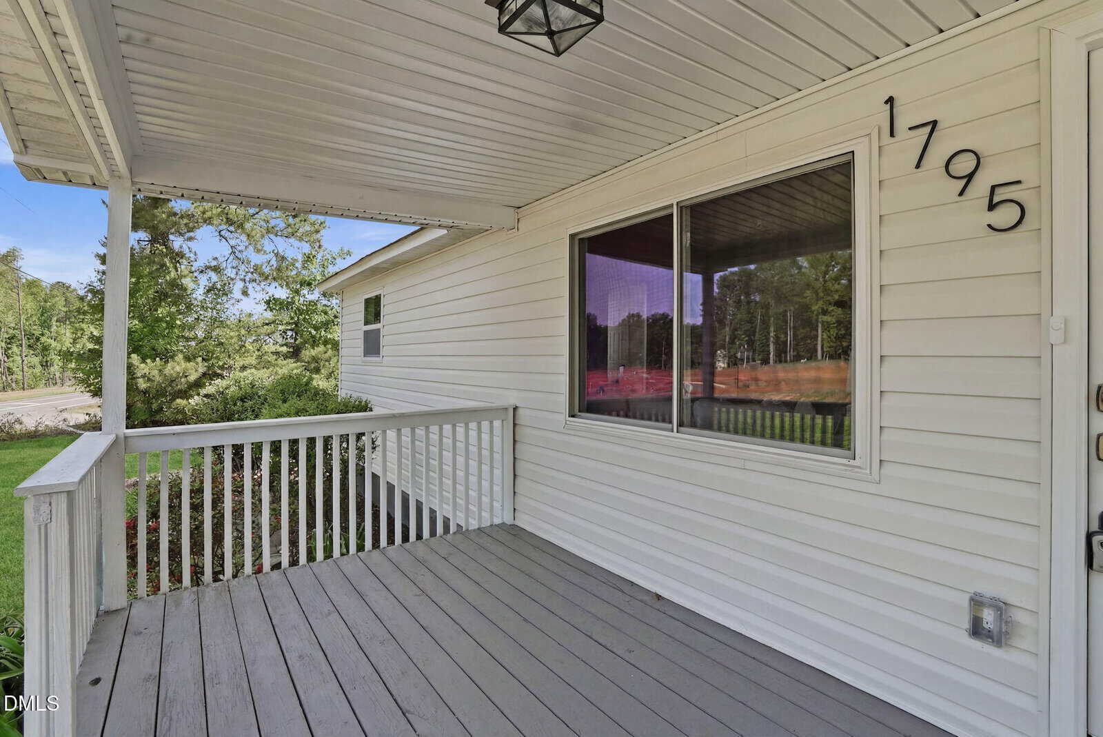 A covered porch with gray flooring and white railing, attached to a light-colored house with large windows and the number 1795 displayed vertically near the door. Green trees and shrubs are visible in the background.