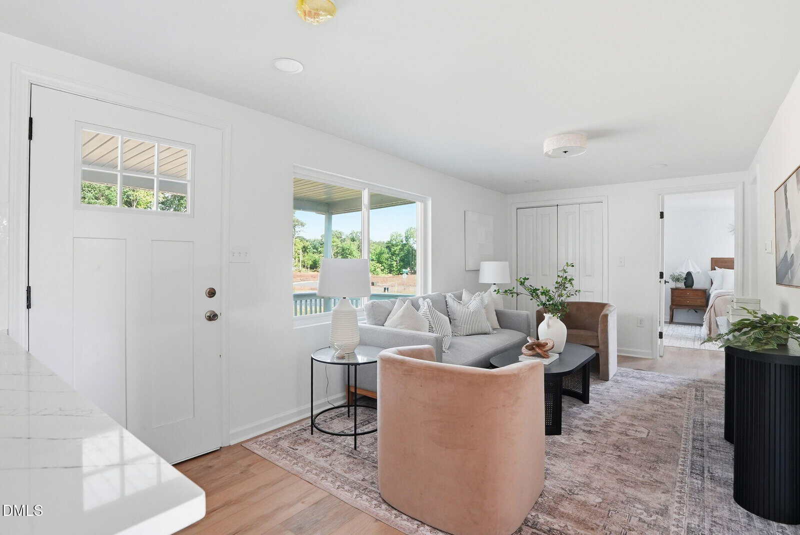 Bright living room with white walls, large windows, a white front door, light wood floors, neutral furniture, a gray sofa, tan chairs, a rug, and plants. A bedroom is visible through an open door in the background.