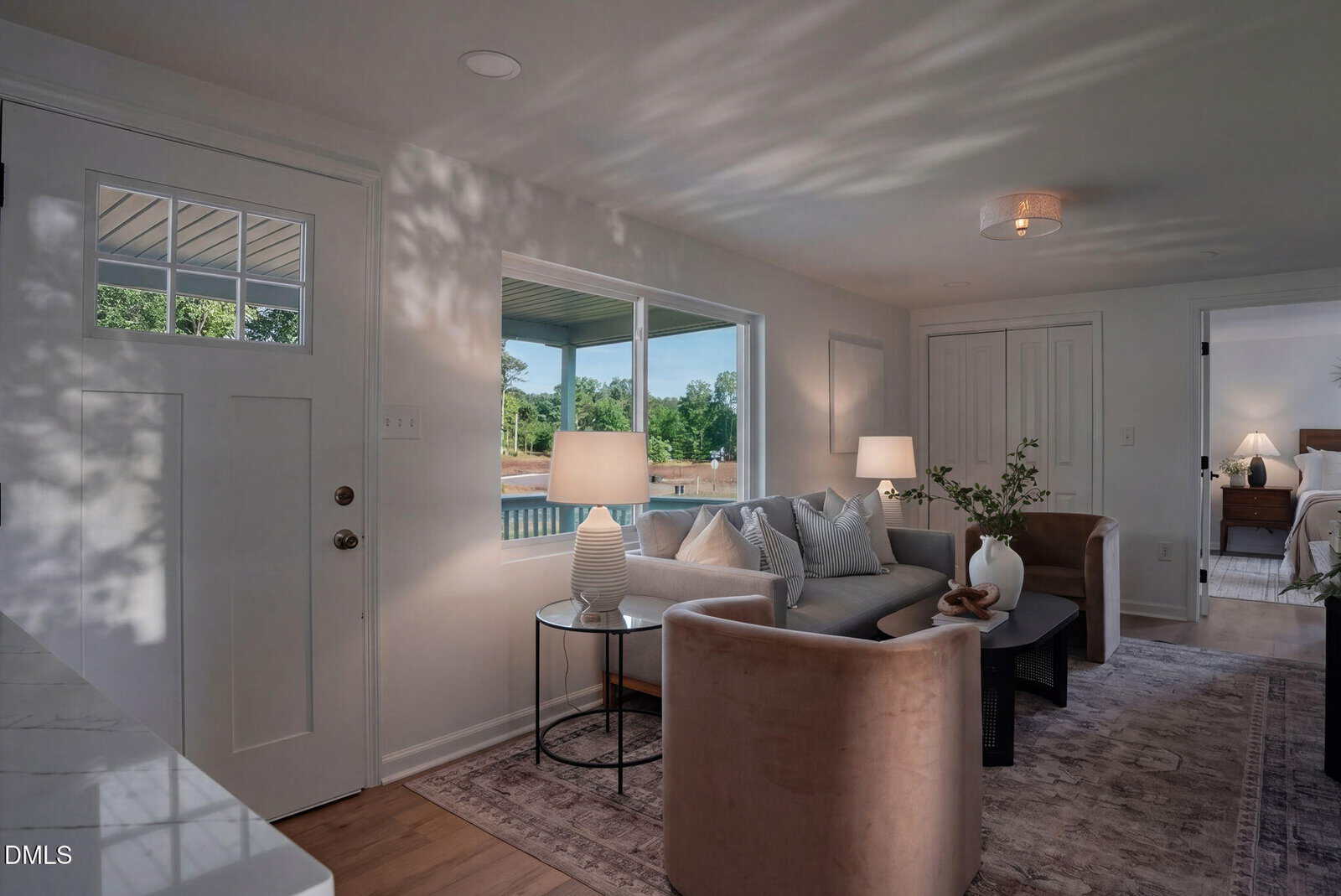 Bright living room with two beige chairs, a gray sofa, a round glass side table, and a large rug. Sunlight streams through windows, showing a porch and green yard. Bedroom visible through open door.