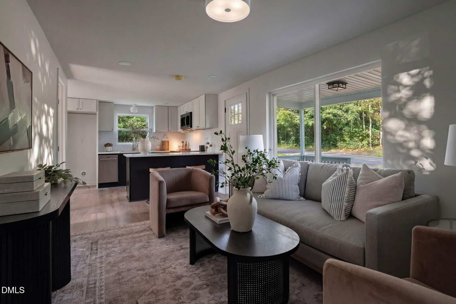 Modern living room with neutral tones, a gray sofa, beige armchair, black coffee table, and large windows letting in natural light. Open kitchen with white cabinets and black island in the background.