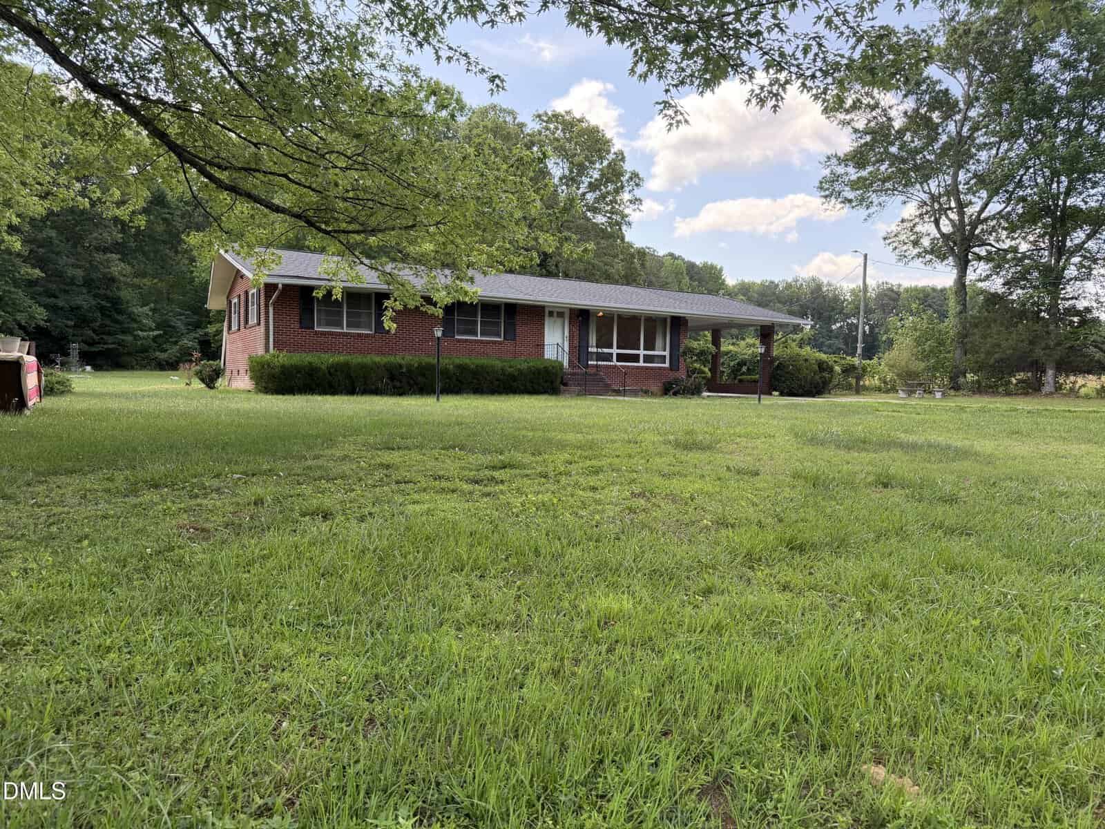 A single-story brick house at 4509 Cannady Mill Road in Oxford sits on a large, grassy yard with trees in the background and partly cloudy sky overhead. Shrubs line the front of the house, and a small porch is visible at the entrance.