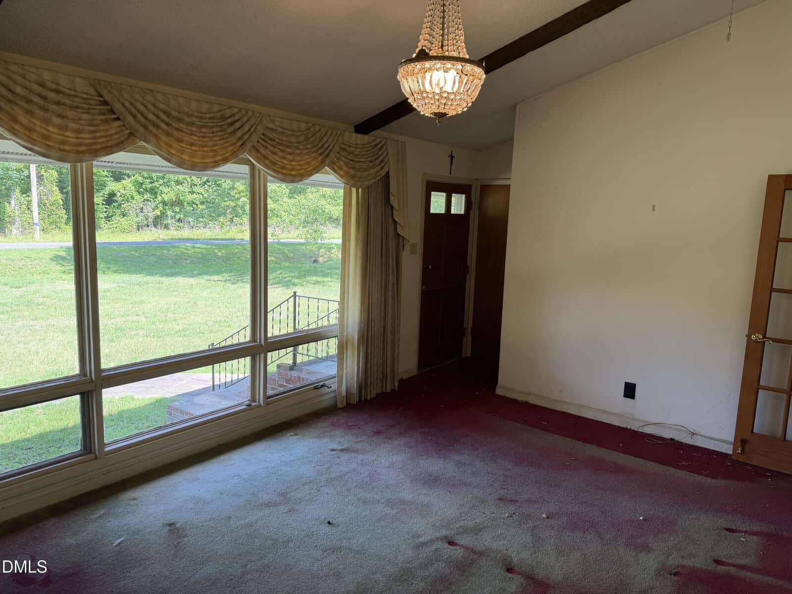 A room with large windows and draped curtains, a red and white faded carpet, a chandelier, a wooden front door, and a white wall with a door leading to another area. Sunlight brightens the space.