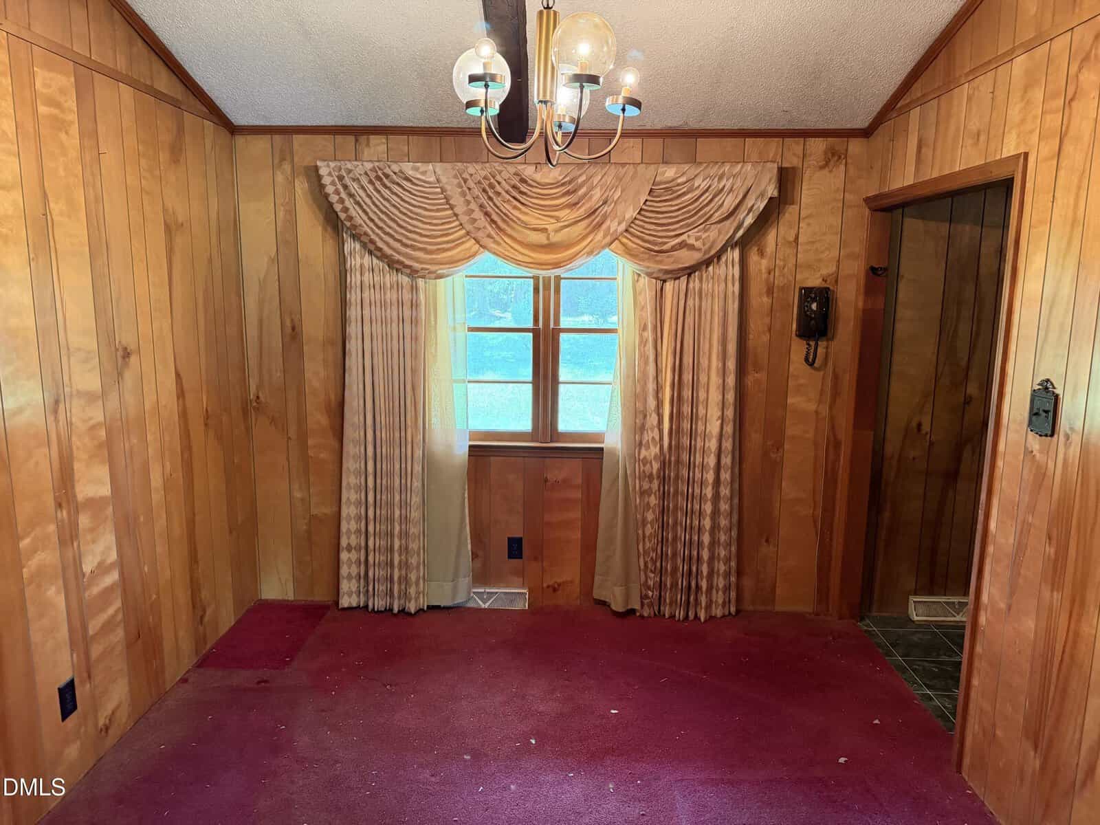 A small room with wood-paneled walls, a red carpet, a window with beige curtains and valance, a vintage chandelier, and an old-fashioned rotary phone mounted on the wall.