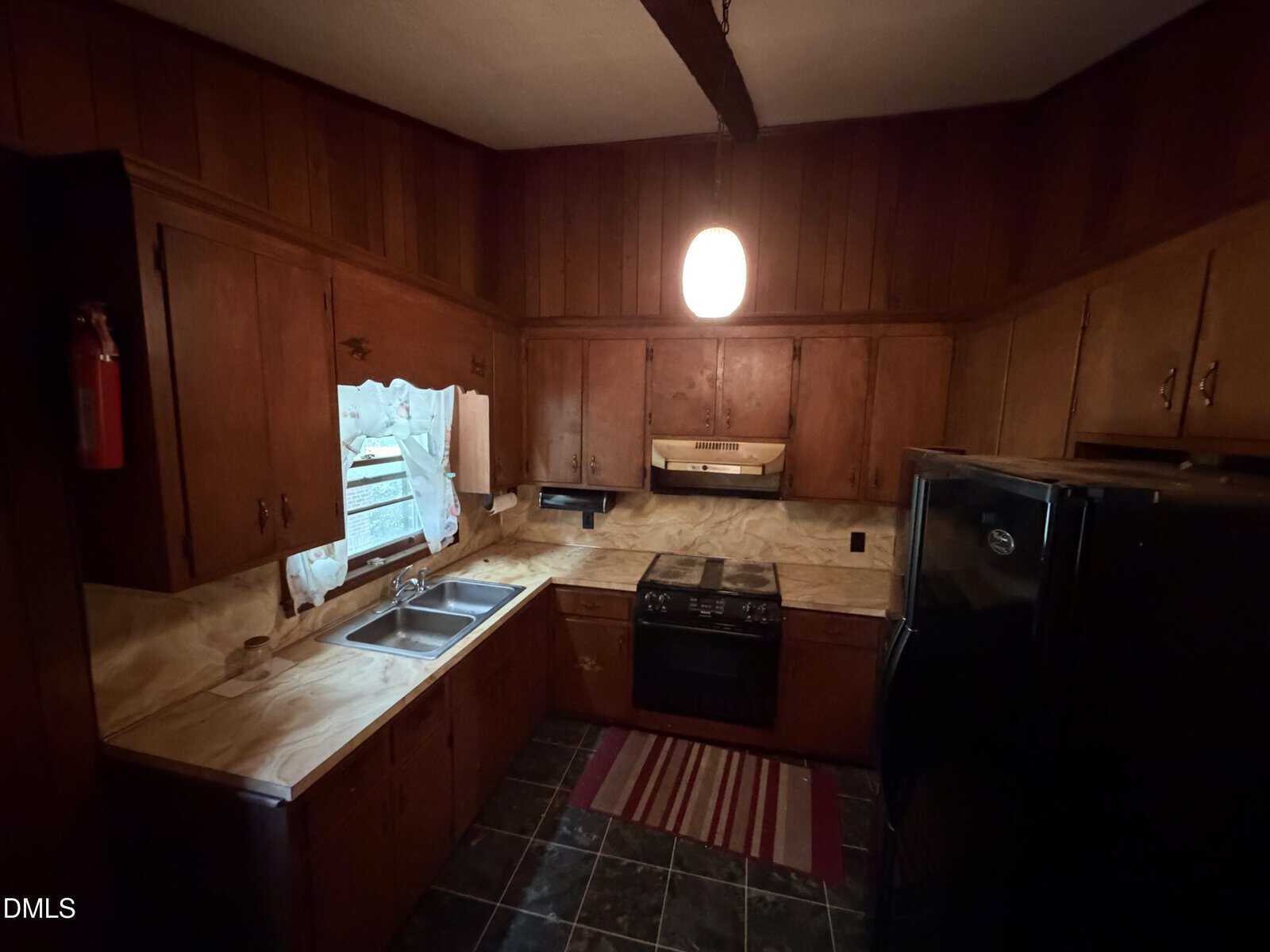 A dimly lit kitchen with dark wood cabinets, marble-patterned countertops, a double sink under a window with curtains, a black stove, black refrigerator, a striped rug, and dark tile flooring. A single ceiling light is on.