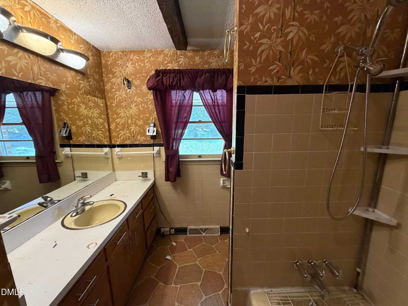 Bathroom with brown floral wallpaper, a beige sink and counter, mirror, two windows with purple curtains, tan tiled shower with handheld showerhead, and hexagonal floor tiles.