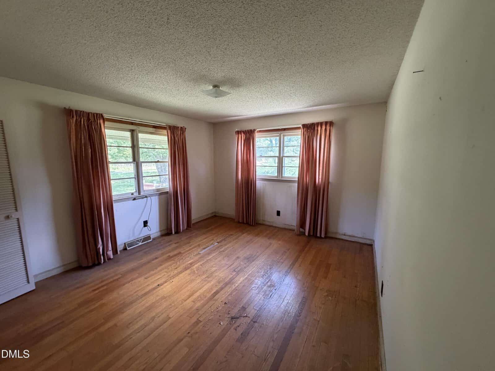 Empty room with hardwood floors, two windows with pink curtains, white walls, and a ceiling light. An air conditioning unit is installed under one window. The room appears spacious and well-lit by natural light.