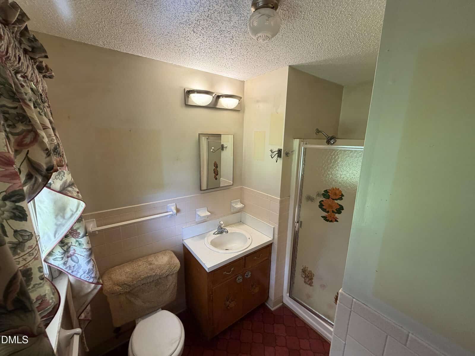 Small bathroom with a sink, vanity, toilet with a fuzzy cover, floral shower door, and patterned curtains. Red flooring and beige walls, with a light fixture and mirror above the sink.