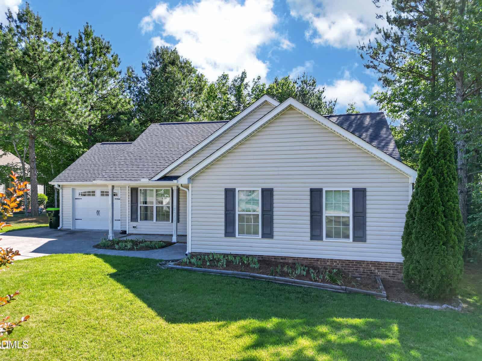 Single-story house with light-colored siding, dark shutters, and a gabled roof. There is a two-car garage, covered front porch, manicured lawn, and trees surrounding the property under a partly cloudy sky.