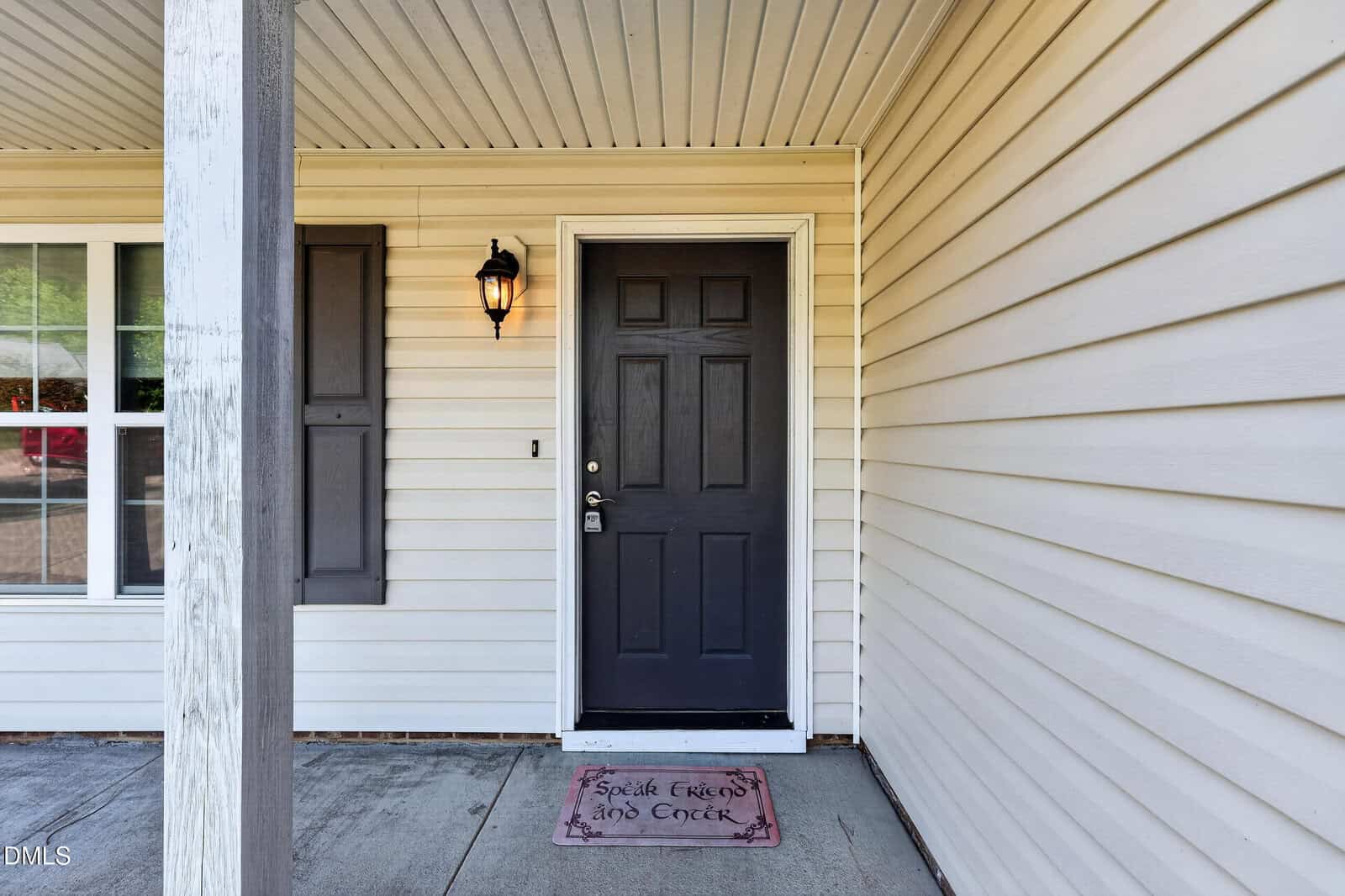 A front porch with beige siding, a black door, a window with closed shutters, a wall-mounted lantern, and a doormat that says "Good Friends and Cheer" on concrete.